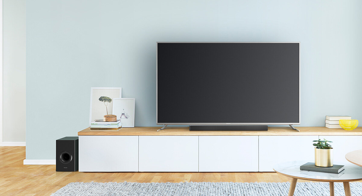 Na obrazie znajduje się nowoczesny salon z dużym telewizorem na białej szafce, obok którego stoi soundbar i subwoofer. Na szafce znajdują się dekoracje, a na podłodze leży dywan.