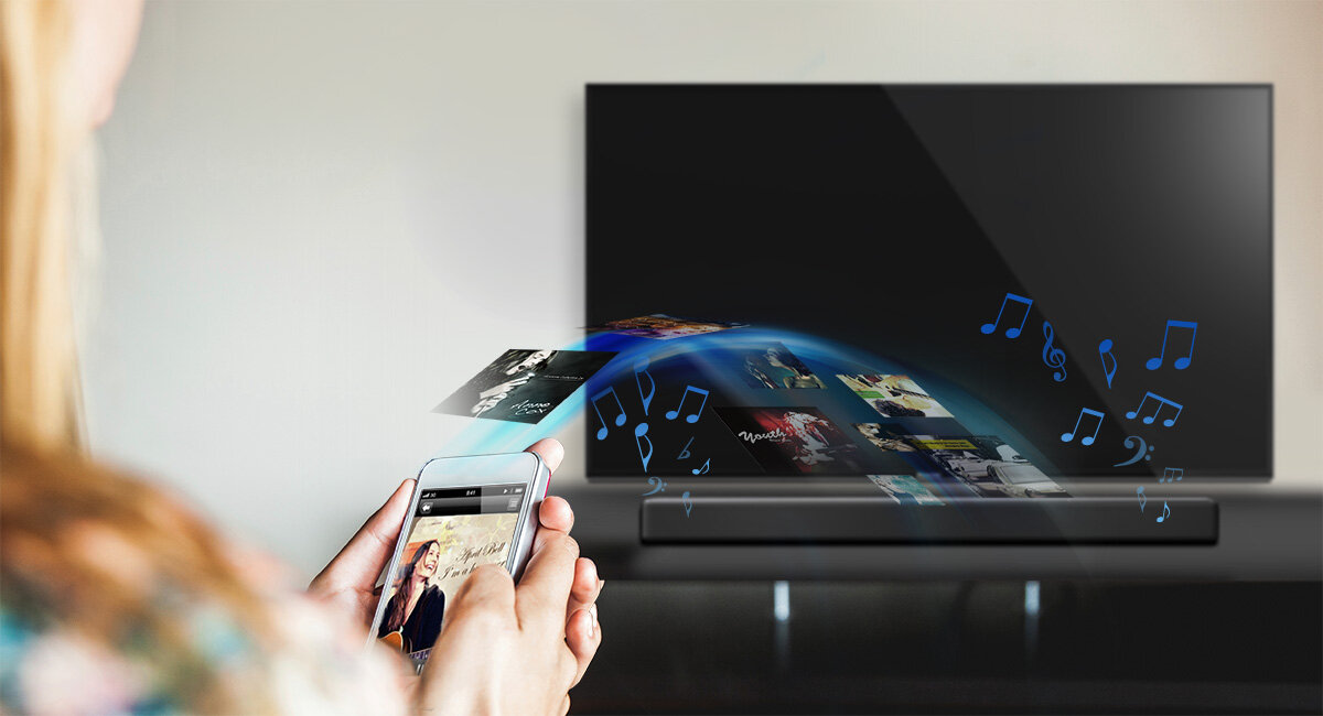Osoba trzymająca smartfon przed telewizorem, z którego wydobywają się kolorowe nuty i obrazy, sugerujące przesyłanie multimediów. Przed telewizorem znajduje się soundbar.