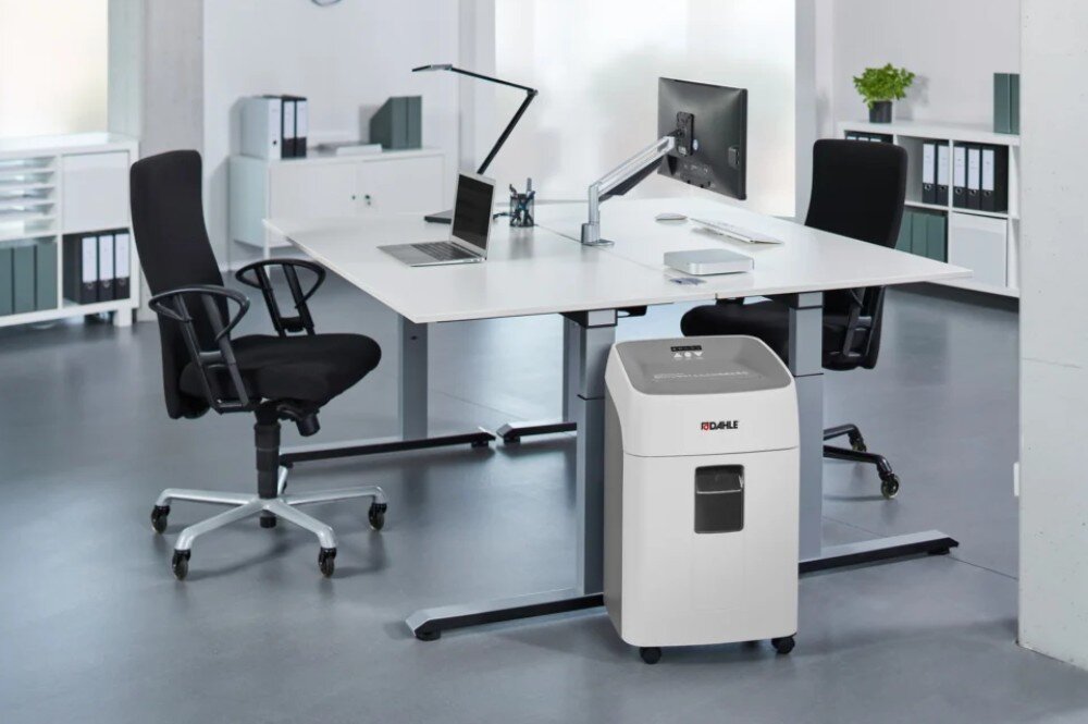 Niszczarka DAHLE Deskside 404P Niszczarka stojąca na kółkach obok nowoczesnego podwójnego biurka z laptopem i monitorem w przestrzeni biurowej