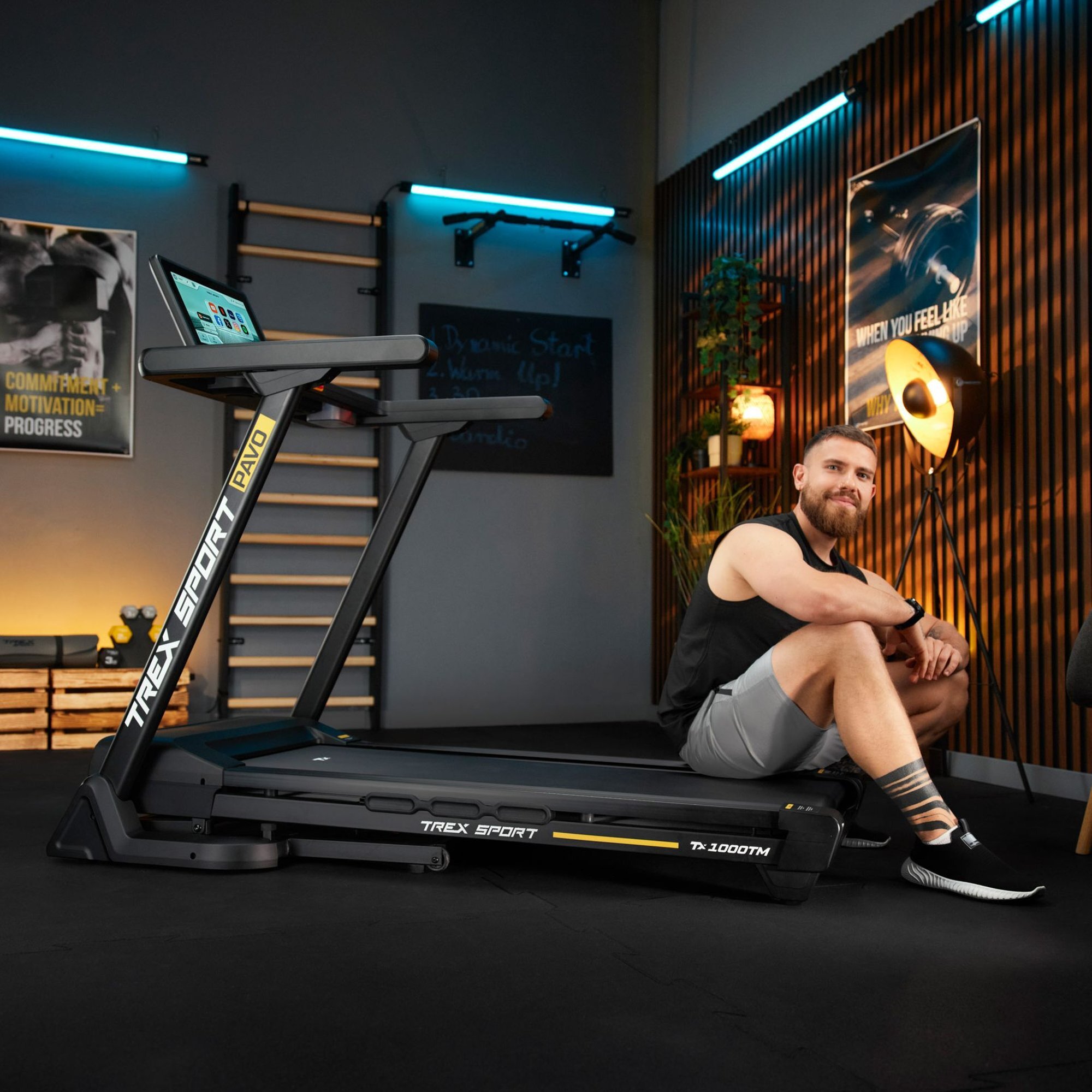 Na obrazie znajduje się bieżnia elektryczna TREX SPORT PAVO z ekranem dotykowym, obok której siedzi mężczyzna w stroju sportowym. W tle widoczne są plakaty i elementy wyposażenia siłowni.