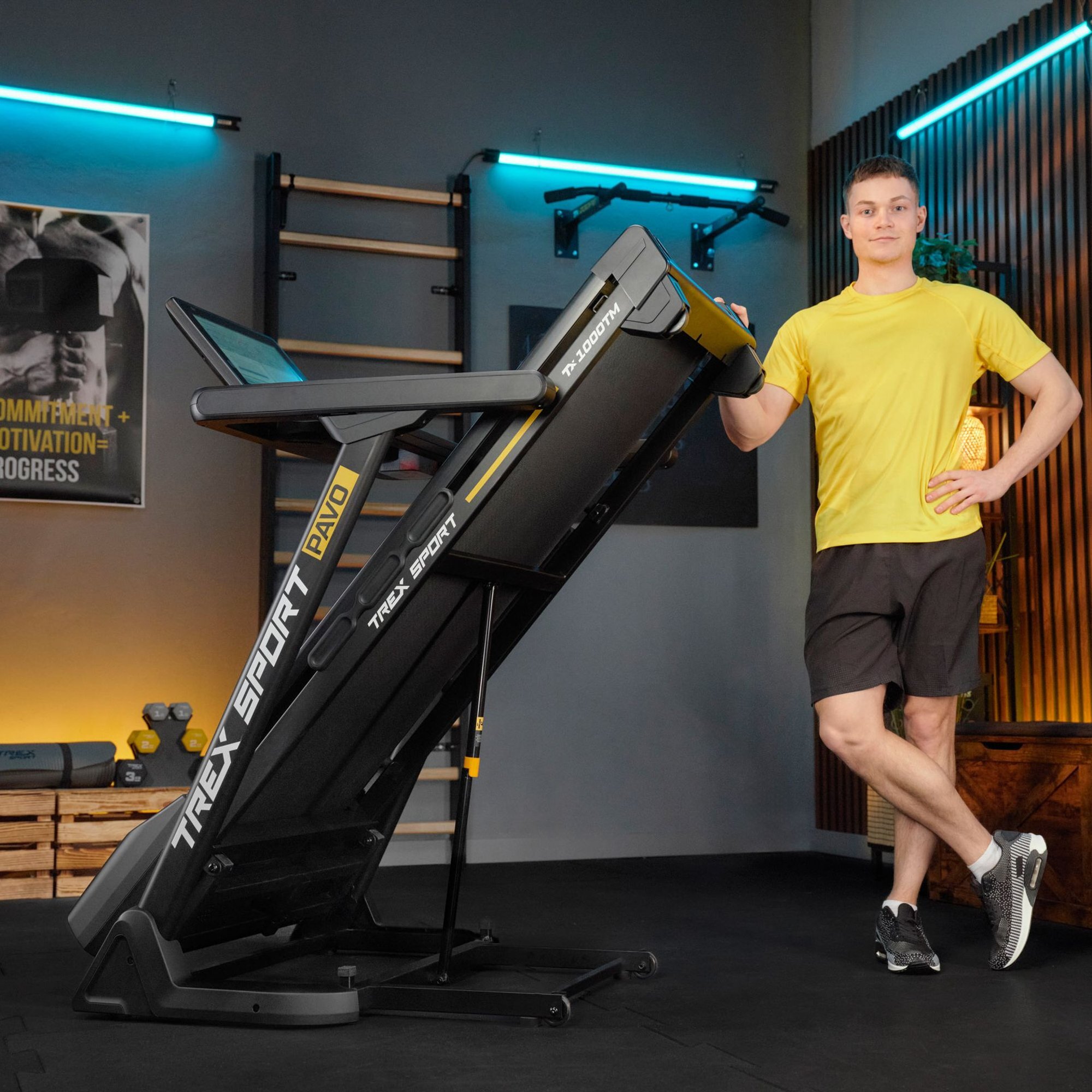 Bieżnia elektryczna TREX SPORT TX-1000TM PAVO SMART jest złożona i ustawiona pionowo. Obok bieżni stoi osoba w żółtej koszulce i czarnych szortach.