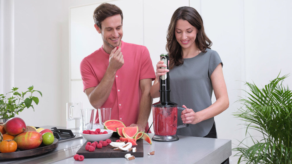 Dwie osoby przygotowują smoothie przy użyciu blendera ręcznego, otoczonego owocami takimi jak arbuz i maliny. W tle widoczne są rośliny doniczkowe i jasne wnętrze kuchni.