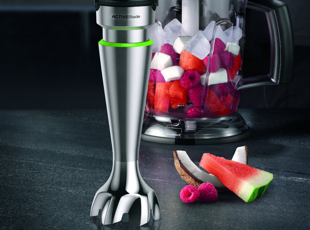 Na obrazie widoczny jest metalowy blender z napisem \'ACTIVEBlade\' oraz pojemnik z pokrojonymi owocami i lodem. Obok leżą kawałki arbuza, kokosa i maliny.