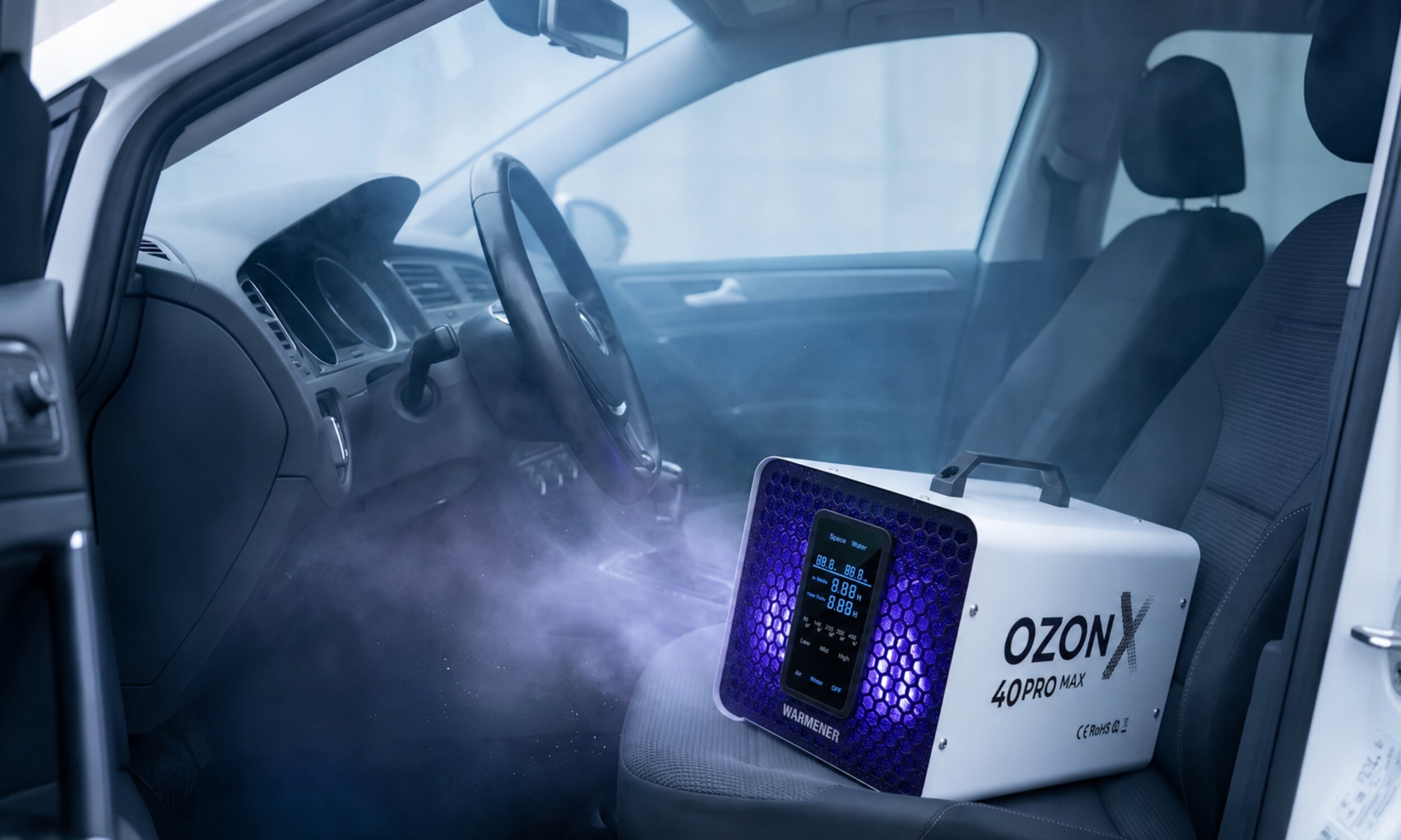 Wnętrze samochodu z urządzeniem oznaczonym jako \'OZON X 40PRO MAX\' i logo \'WARMENER\', emitującym mgłę. Urządzenie znajduje się na siedzeniu pasażera.