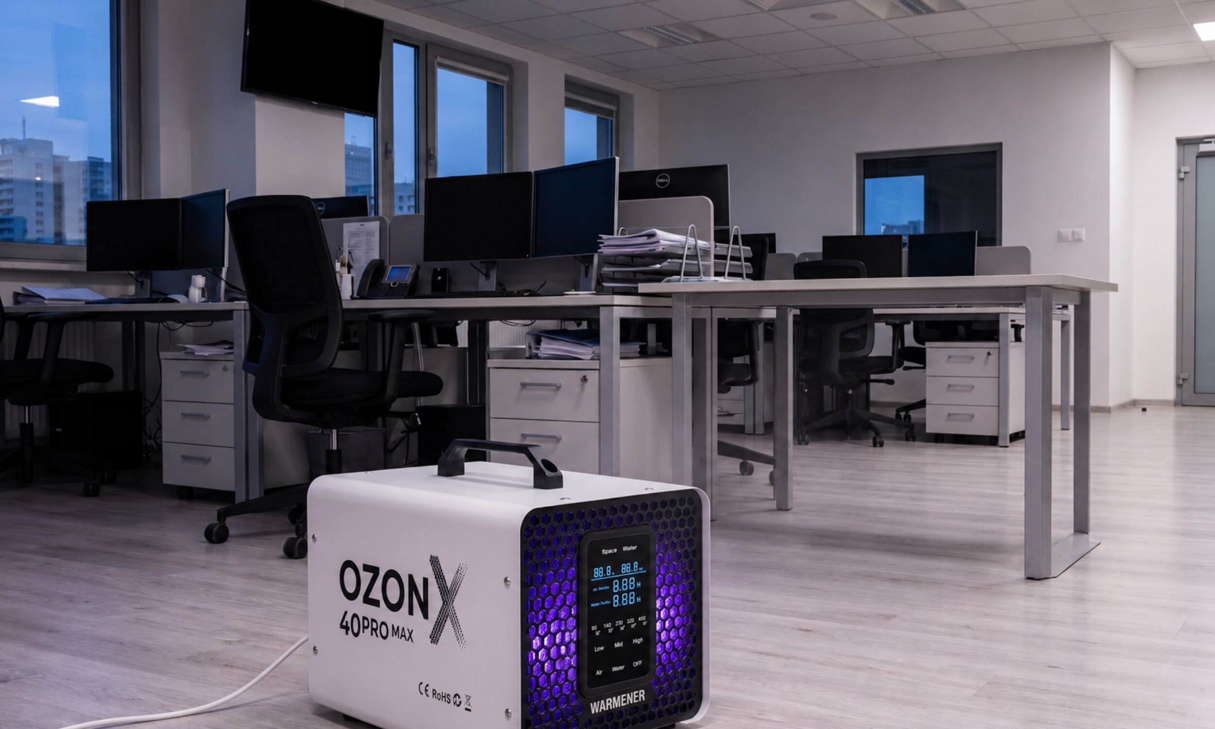 W biurze na podłodze stoi generator ozonu z napisem \'OzonX 40Pro Max\' i logo \'WARMENER\'. W tle widać biurka z komputerami i krzesłami.