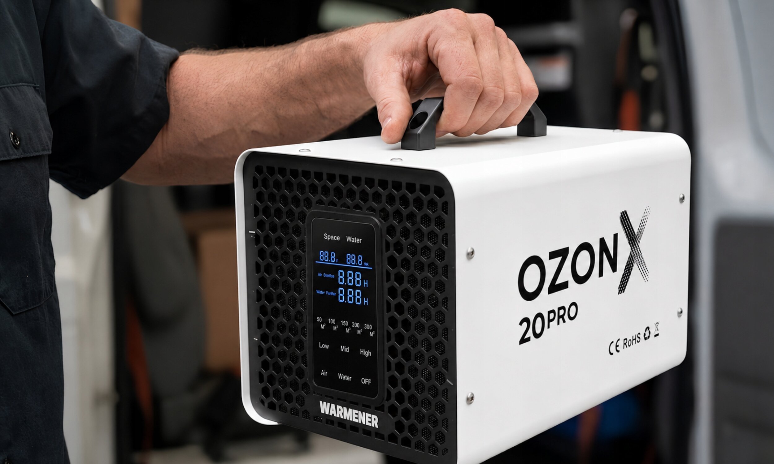 Osoba trzyma białe urządzenie z czarnym panelem przednim, na którym widoczny jest wyświetlacz z niebieskimi cyframi. Na boku urządzenia znajduje się napis \'OZON X 20PRO\', a poniżej logo \'WARMENER\'.