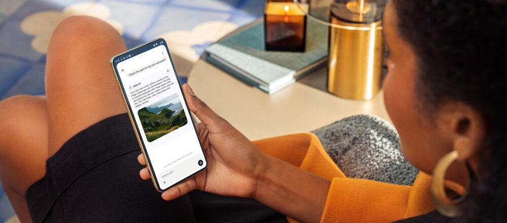 Osoba trzyma smartfon, na ekranie widoczny jest tekst i zdjęcie krajobrazu. W tle znajdują się książki i złoty pojemnik na stole.