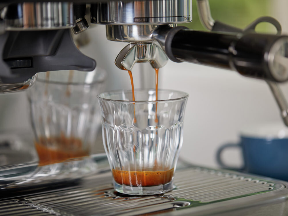 Ekspres do kawy nalewa espresso do szklanki umieszczonej na metalowej tacy ociekowej. W tle widoczna jest rozmyta filiżanka.