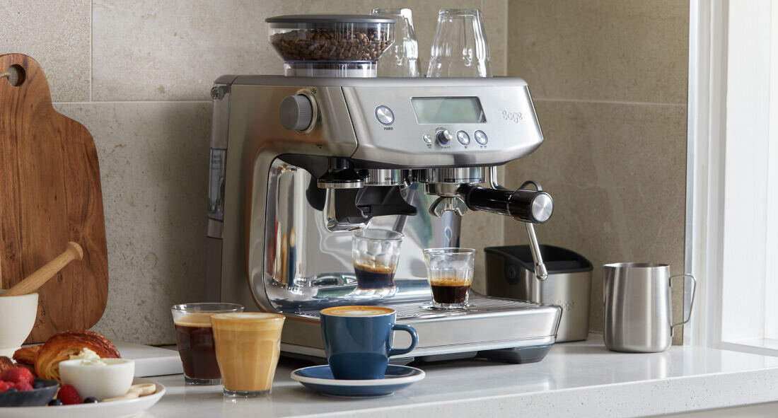 Na blacie kuchennym stoi srebrny ekspres do kawy z wyświetlaczem i dwoma filiżankami espresso. Obok znajdują się filiżanki z kawą, dzbanek na mleko oraz deska z pieczywem i owocami.