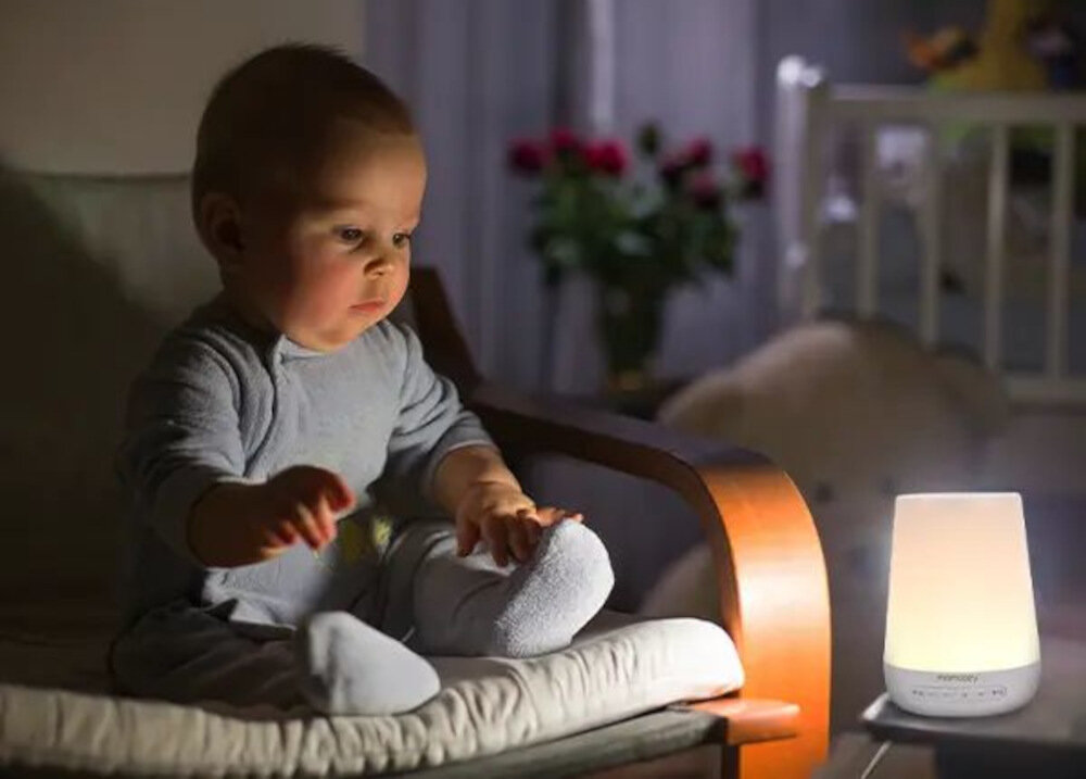 Na zdjęciu widać dziecko siedzące na fotelu oraz lampkę nocną emitującą ciepłe światło. Na lampce widoczny jest napis \'MOMCOZY\'.
