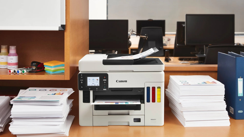 Na biurku stoi urządzenie wielofunkcyjne Canon z widocznym logo, otoczone stosami papieru i materiałami biurowymi. W tle znajdują się monitory komputerowe.