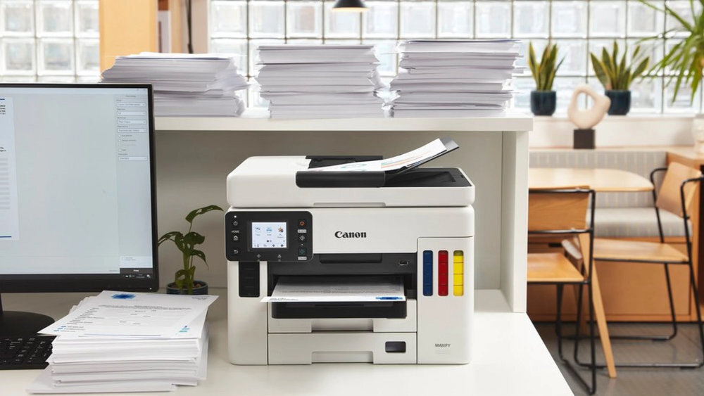 Na biurku znajduje się urządzenie wielofunkcyjne Canon z wyświetlaczem i kolorowymi wskaźnikami poziomu tuszu. W tle widać stosy papieru, monitor oraz rośliny doniczkowe.