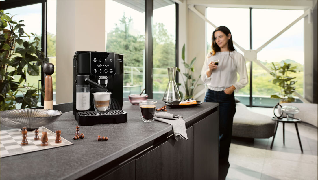 Na blacie kuchennym stoi ekspres do kawy z logo DELONGHI, obok filiżanki z kawą i akcesoria kuchenne. W tle widać osobę trzymającą filiżankę, a za nią duże okna z widokiem na zieleń.