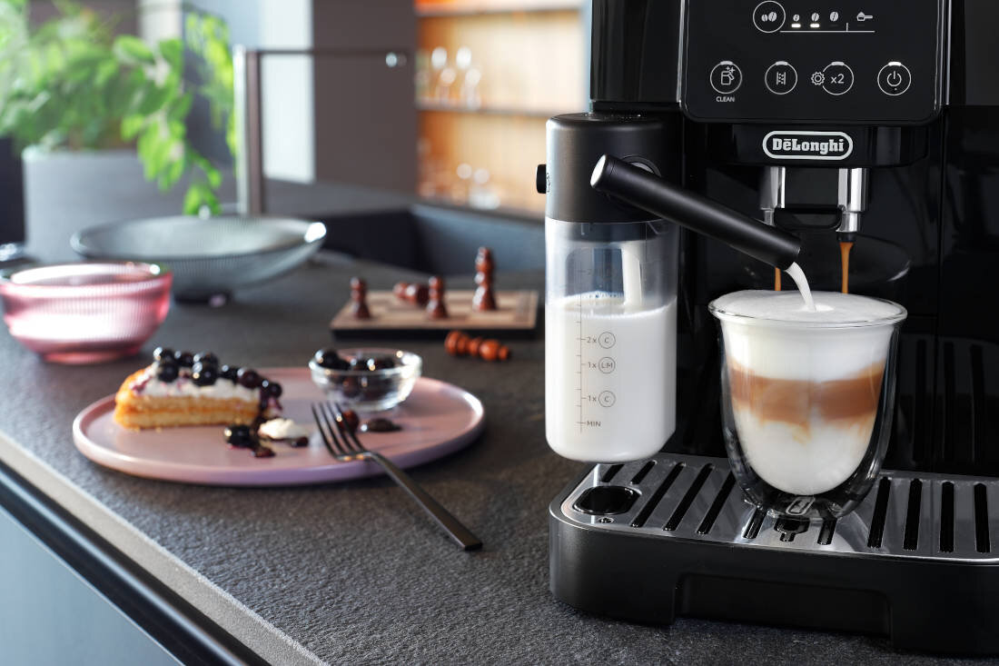 Na blacie kuchennym stoi ekspres do kawy z logo \'DeLonghi\', przygotowujący kawę z mlekiem. Obok znajduje się talerz z kawałkiem ciasta i widelcem, a w tle widać szachy.