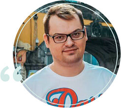 Zdjęcie przedstawia mężczyznę w okularach, ubranego w biały t-shirt z kolorowym nadrukiem. W tle widoczne są elementy architektury miejskiej.