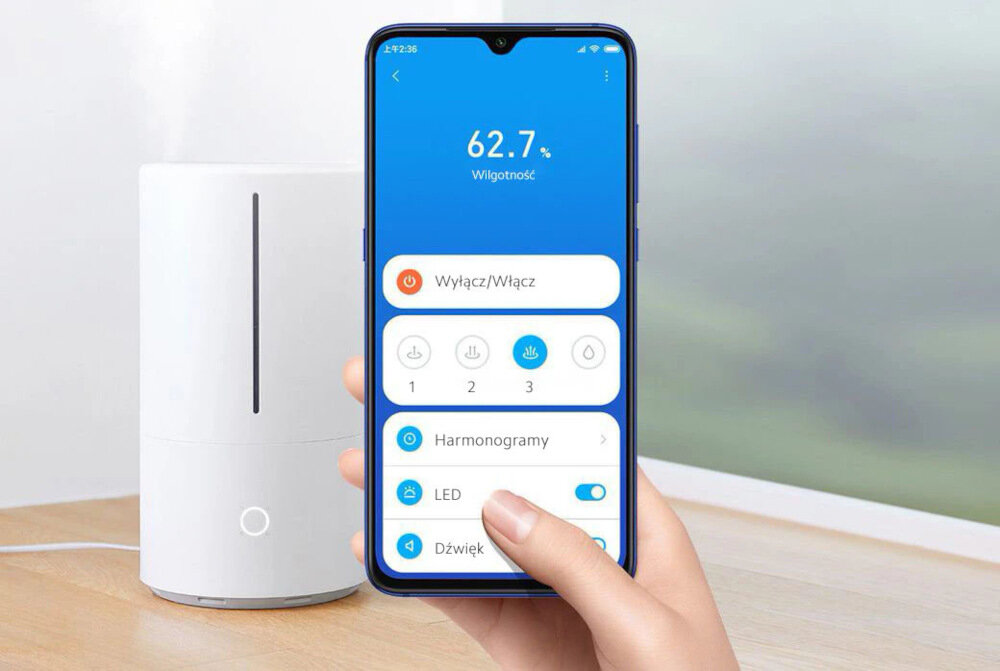 Na zdjęciu widać biały nawilżacz ultradźwiękowy oraz dłoń trzymającą smartfon z aplikacją pokazującą wilgotność 62.7% i opcje sterowania urządzeniem. W tle znajduje się rozmyty widok za oknem.