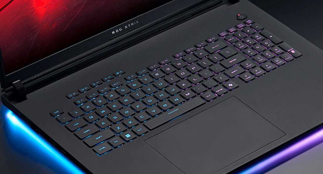 Laptop z podświetlaną klawiaturą i napisem \'ROG STRIX\' nad klawiaturą. Obudowa ma ciemny kolor, a wokół laptopa widoczne są kolorowe efekty świetlne.