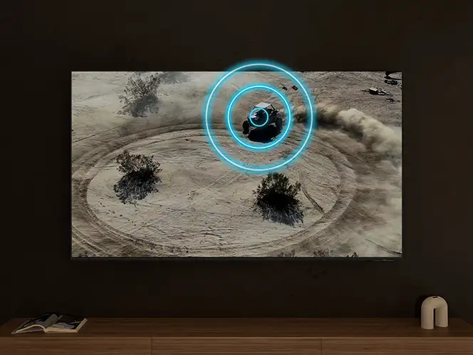 Telewizor wyświetla obraz pustynnego krajobrazu z krzewami i pojazdem, wokół którego widoczne są niebieskie okręgi. Na drewnianej półce pod telewizorem leży książka i stoi biały głośnik.