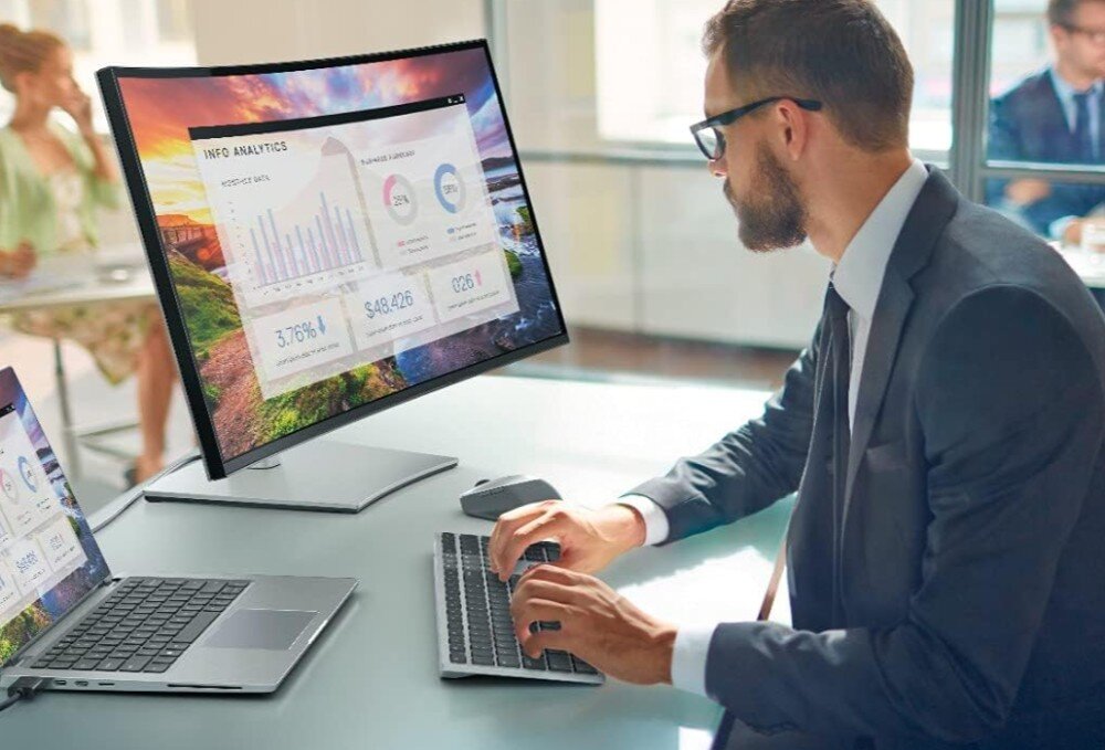 Mężczyzna w garniturze pracuje przy biurku z dwoma laptopami i zakrzywionym monitorem, na którym wyświetlane są wykresy i dane analityczne. Obok znajduje się klawiatura i myszka, które mogą być częścią zestawu DELL KM7321W.