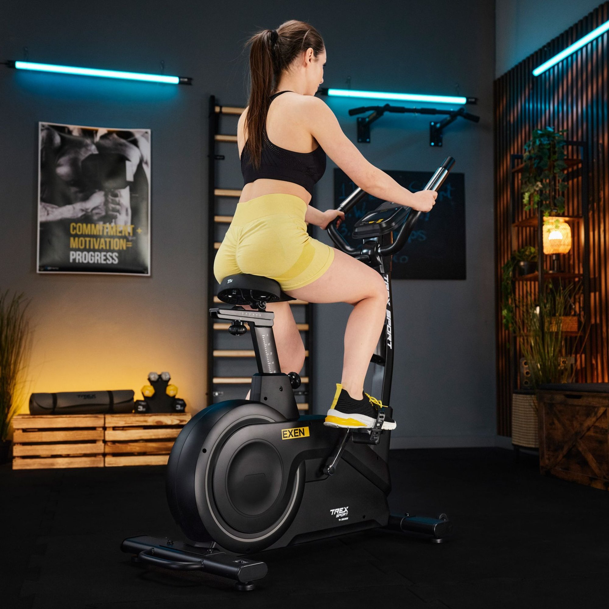Osoba ćwiczy na czarnym rowerze stacjonarnym elektryczno-magnetycznym EXEN TX-950EB w pomieszczeniu z plakatem \'COMMITMENT. MOTIVATION. PROGRESS\'. W tle widoczne są elementy dekoracyjne i sprzęt do ćwiczeń.