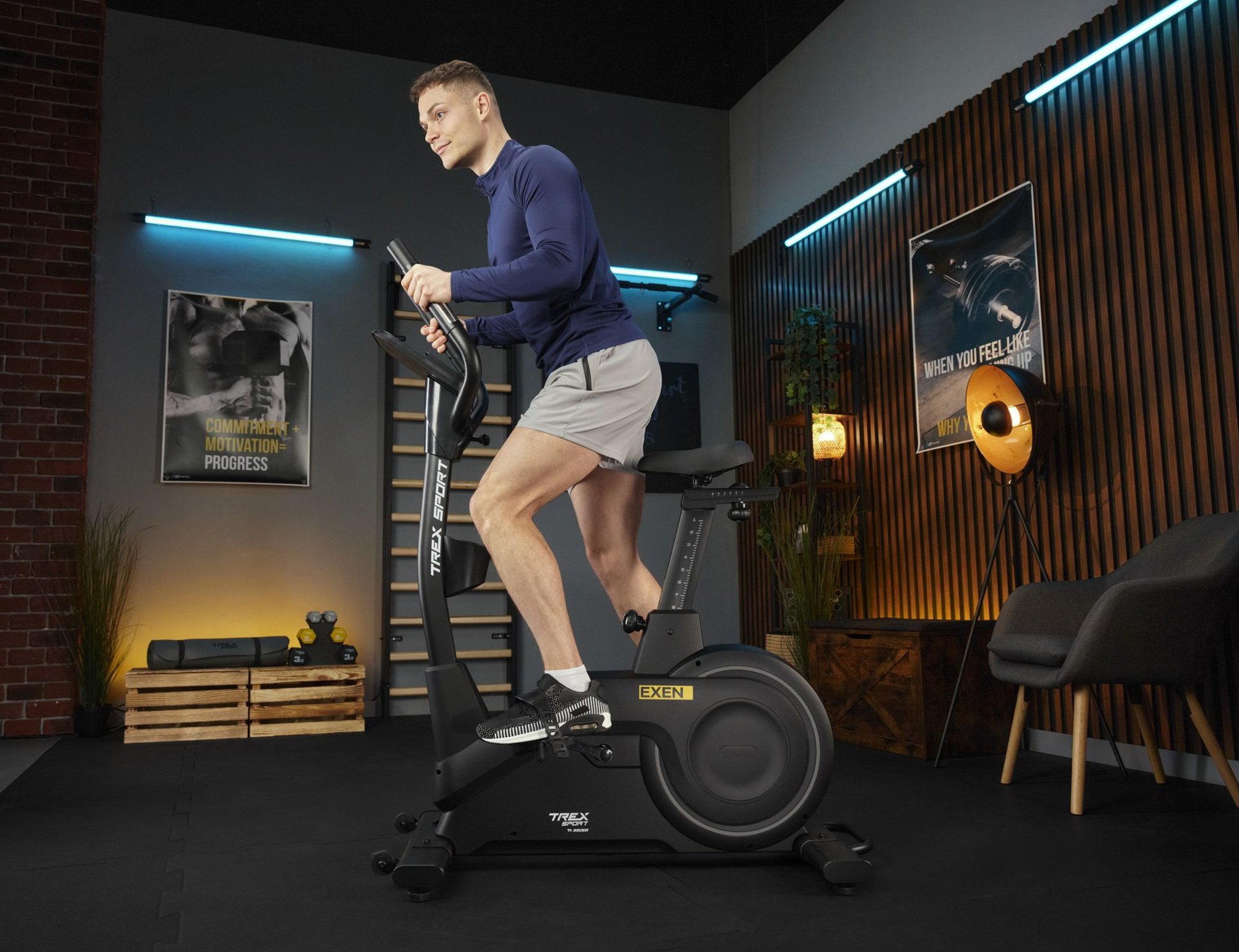 Osoba ćwiczy na czarnym rowerze stacjonarnym EXEN TX-950EB w nowoczesnym wnętrzu z plakatami na ścianach. Na rowerze widoczne są napisy \'TREX SPORT\' i \'EXEN\'.