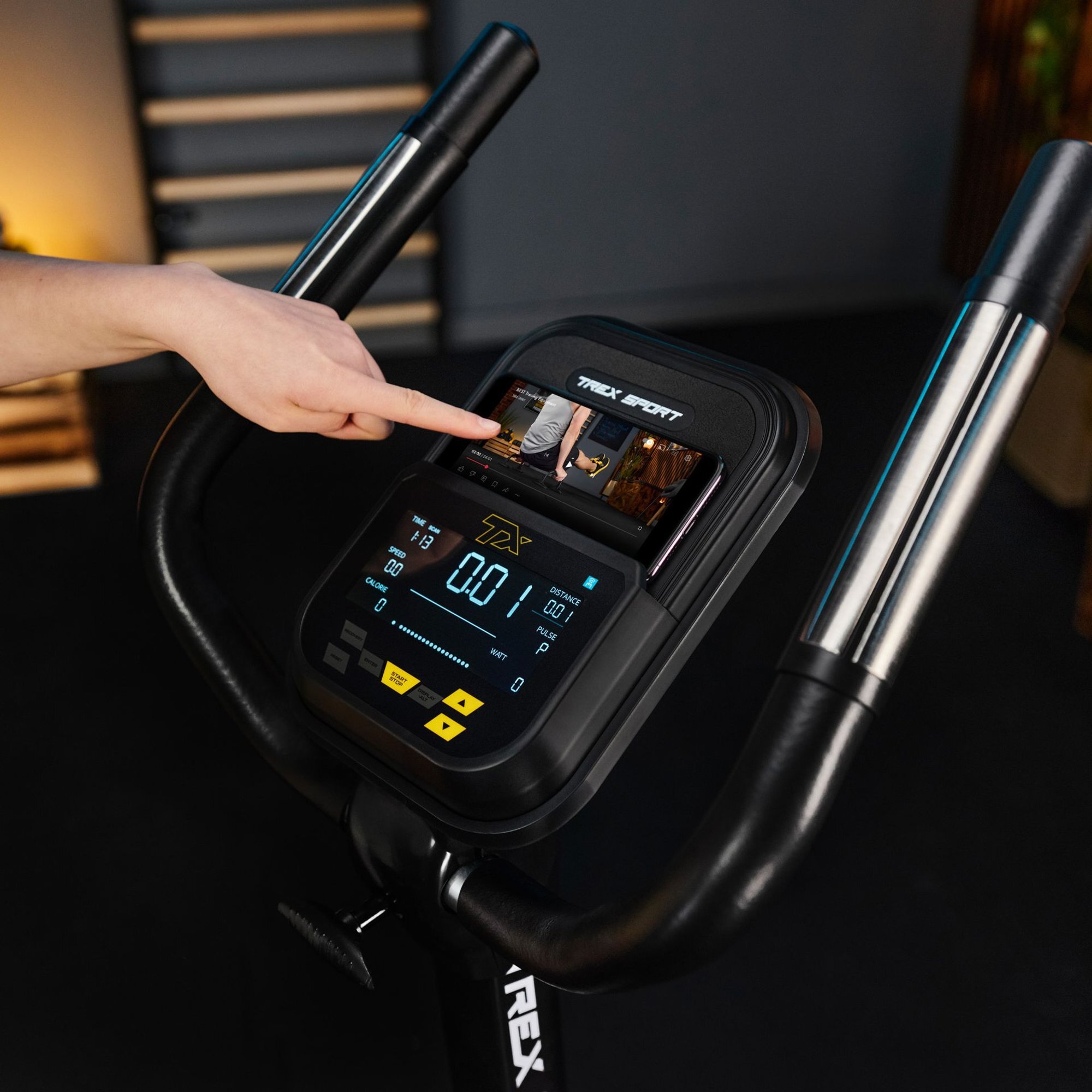 Osoba dotyka ekranu panelu sterującego roweru stacjonarnego z wyświetlaczem pokazującym dane treningowe. Na panelu widoczne jest logo \'TREX SPORT\'.