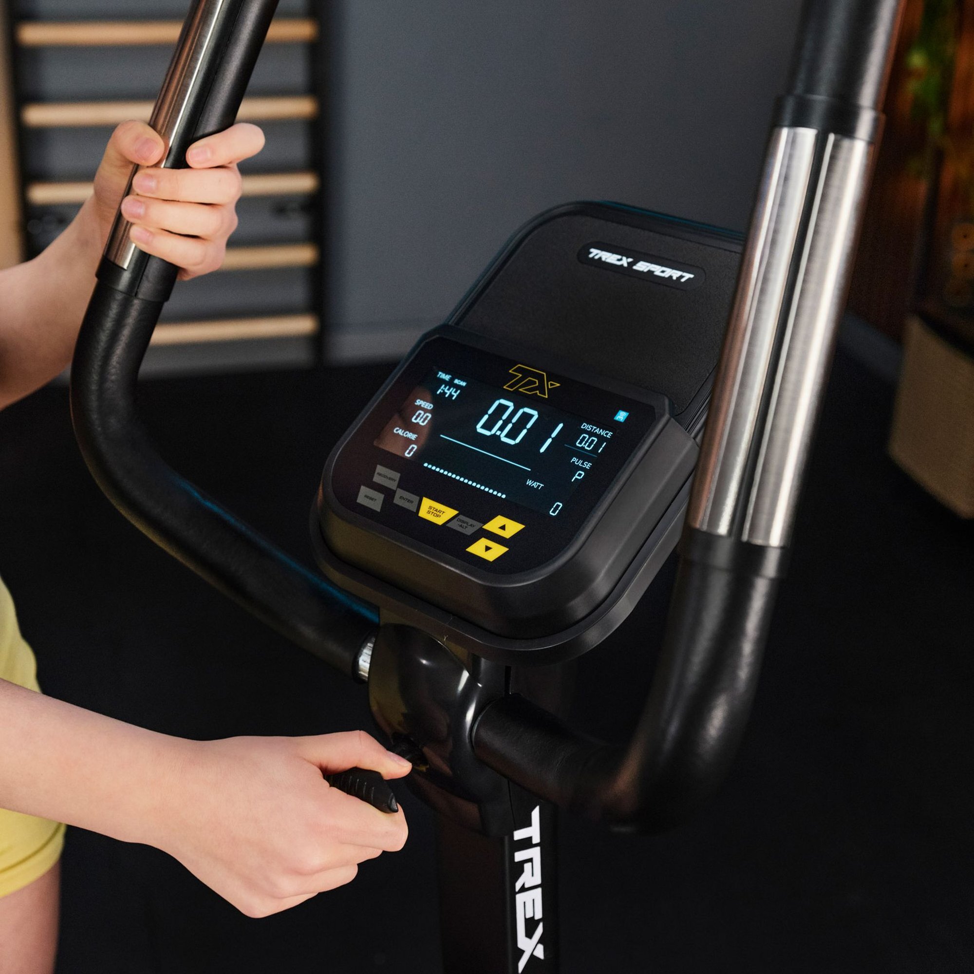 Osoba trzyma kierownicę roweru stacjonarnego z wyświetlaczem pokazującym dane treningowe. Na wyświetlaczu widoczne są wartości: 144, 0.01, 0 oraz logo TREX SPORT.