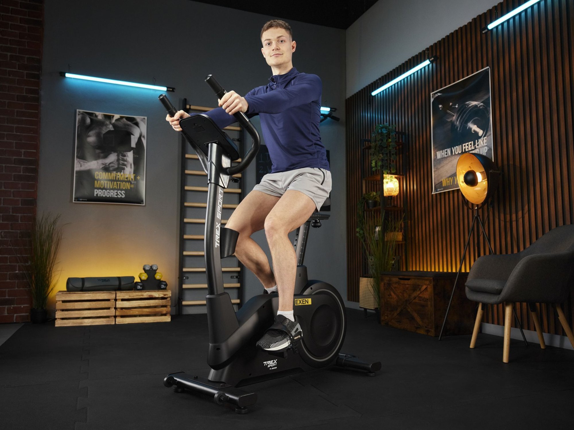 Osoba ćwiczy na czarnym rowerze stacjonarnym elektryczno-magnetycznym EXEN TX-950EB w nowoczesnym wnętrzu. Na ścianie widoczne są plakaty z napisami, a w pomieszczeniu znajdują się elementy dekoracyjne i sprzęt do ćwiczeń.