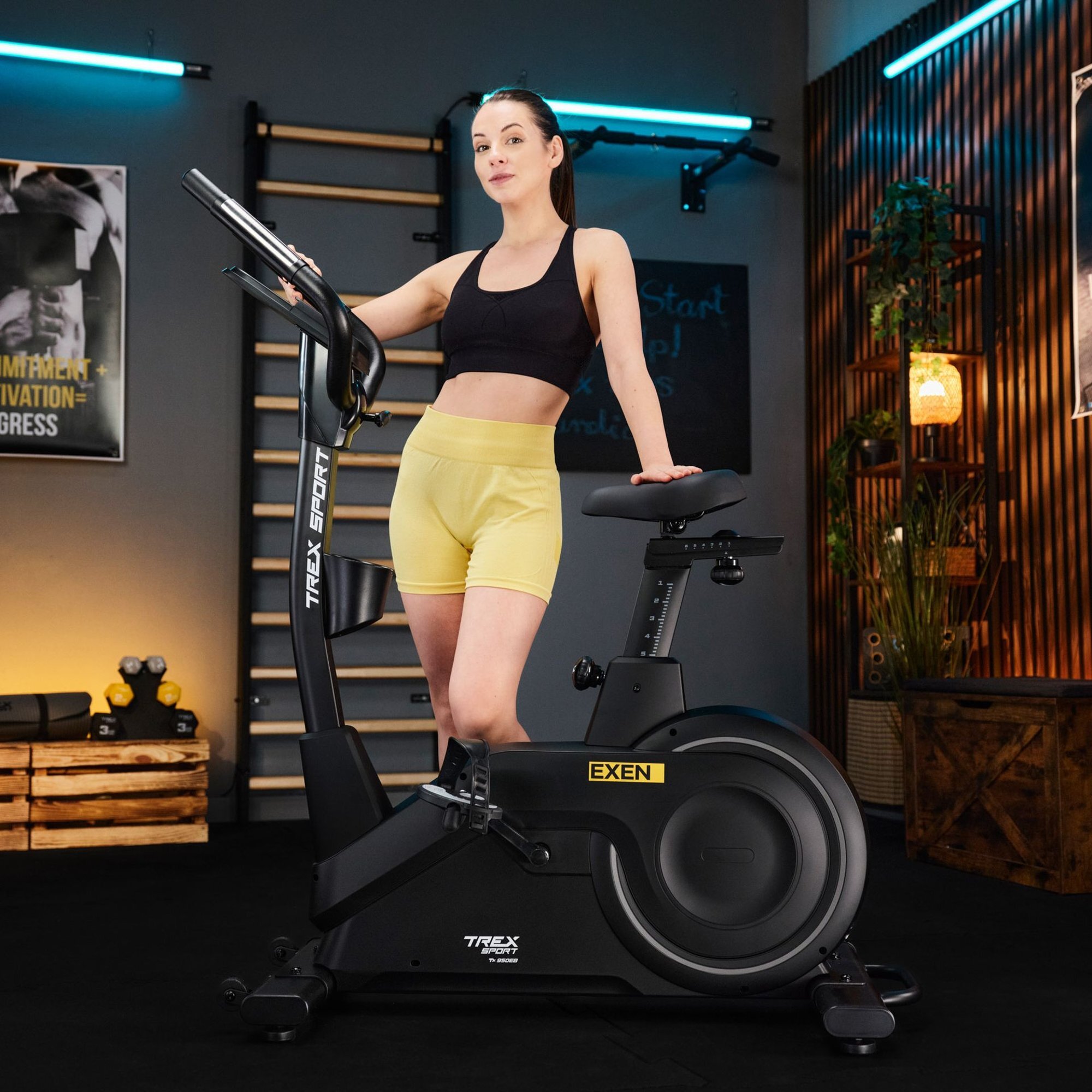 Czarny rower stacjonarny elektryczno-magnetyczny z logo EXEN i TREX SPORT, na którym stoi osoba w żółtych szortach i czarnym topie. W tle widoczne są elementy wyposażenia siłowni, takie jak drabinki i hantle.