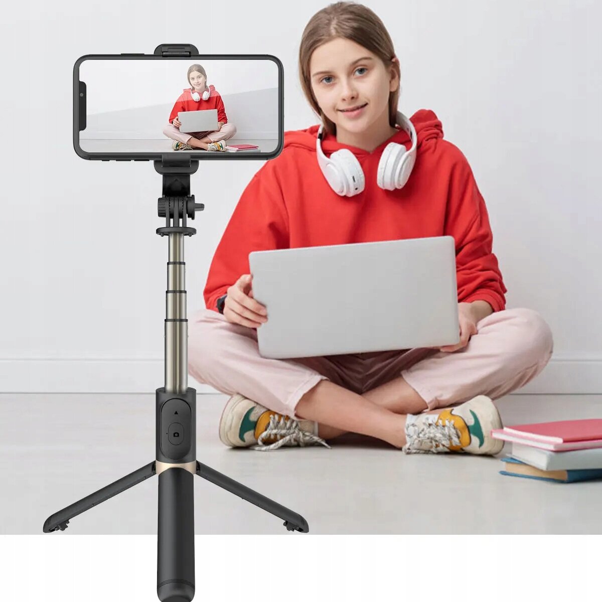 Osoba siedzi na podłodze z laptopem na kolanach, a przed nią stoi kijek selfie z zamocowanym telefonem. Na ekranie telefonu widoczny jest obraz tej samej osoby w tej samej pozycji.