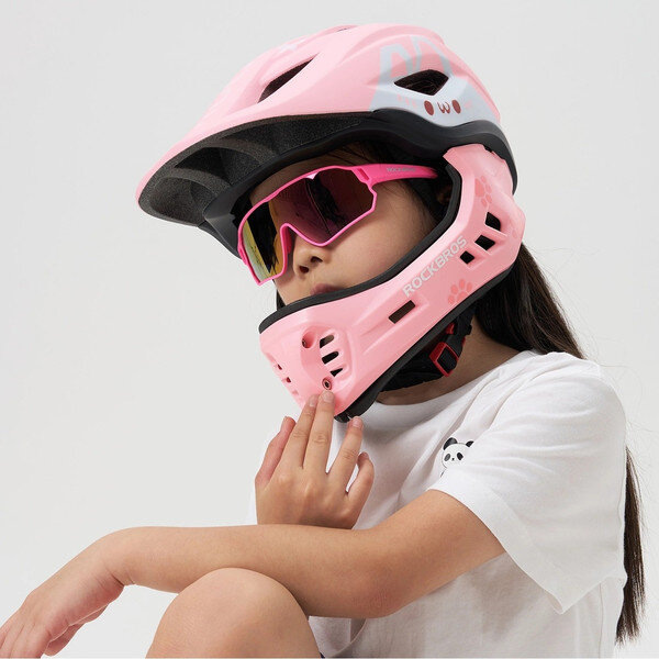 Dziecko nosi różowy kask rowerowy z odpinaną szczęką i różowe okulary przeciwsłoneczne. Na kasku widoczny jest napis \'ROCKBROS\'.