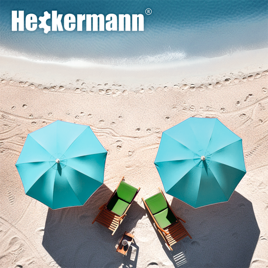 Na plaży znajdują się dwa turkusowe parasole plażowe oraz dwa leżaki z zielonymi siedziskami. W górnej części obrazu widoczne jest logo \'Heckermann\'.
