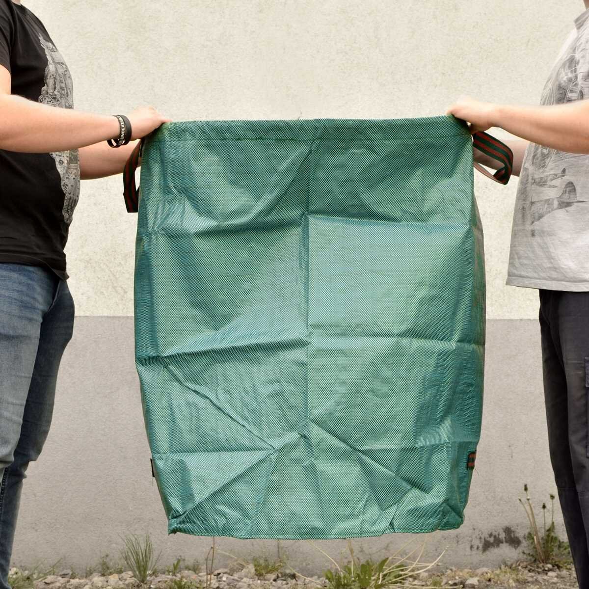Zielona torba ogrodowa Heckermann 330L jest trzymana przez dwie osoby na tle szarej ściany i trawy.