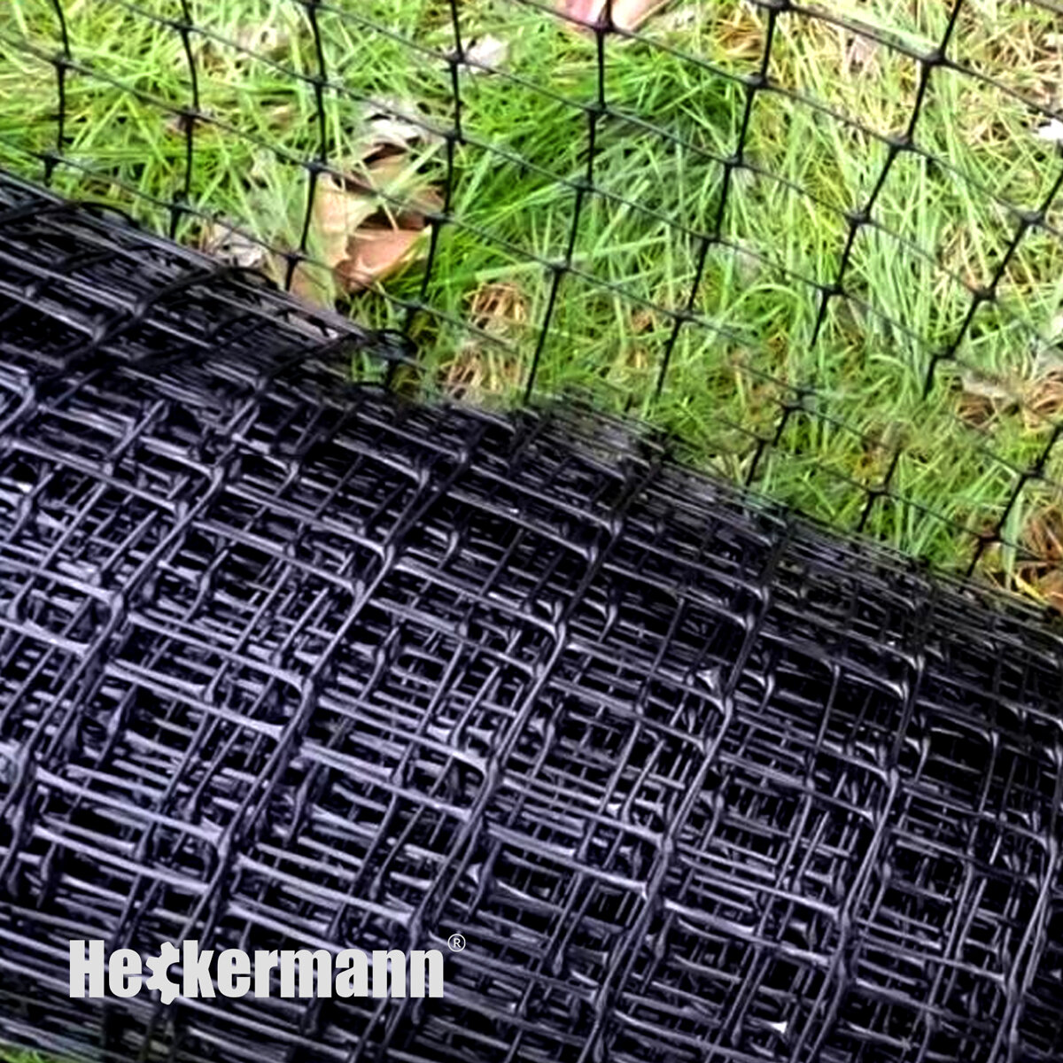 Zdjęcie przedstawia czarną siatkę ogrodową zwiniętą w rolkę, leżącą na trawie. Na siatce widoczne jest logo \'Heckermann\'.