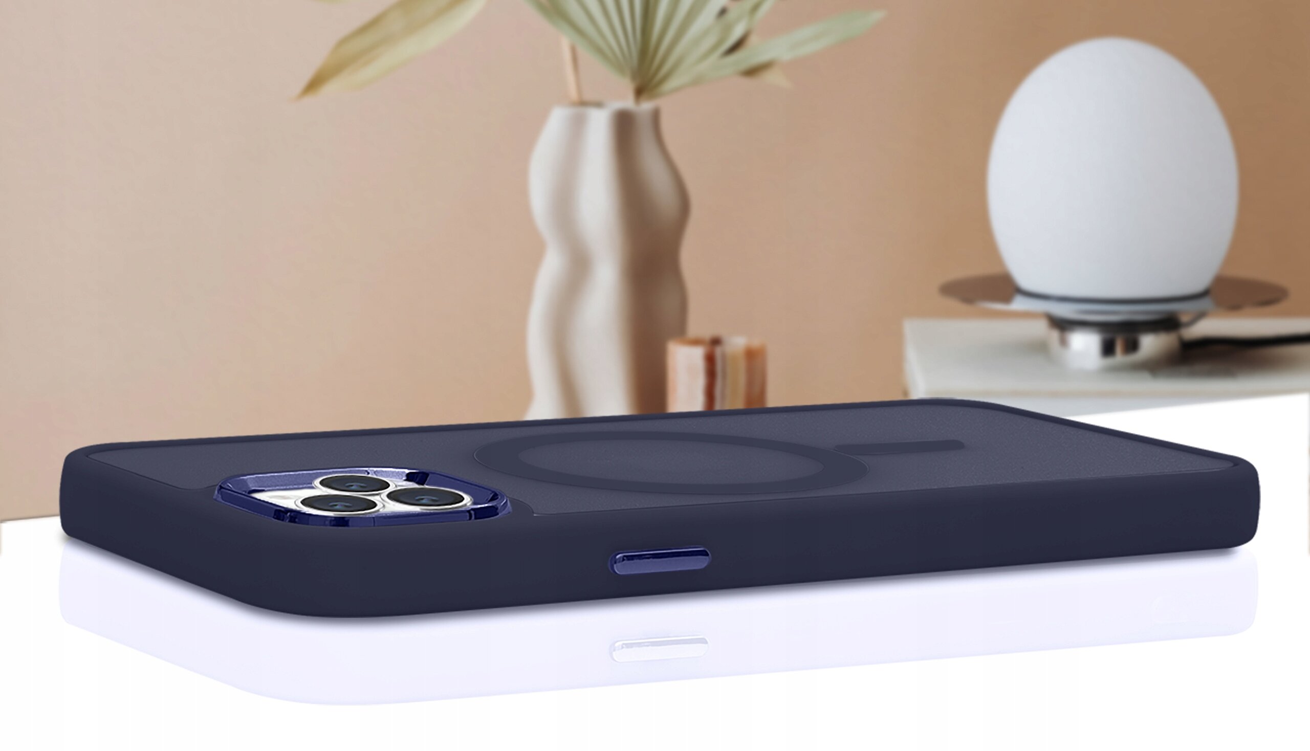Etui do iPhone 15 Pro Max w kolorze ciemnym, leży na białej powierzchni, w tle widoczny jest wazon z rośliną i lampa.