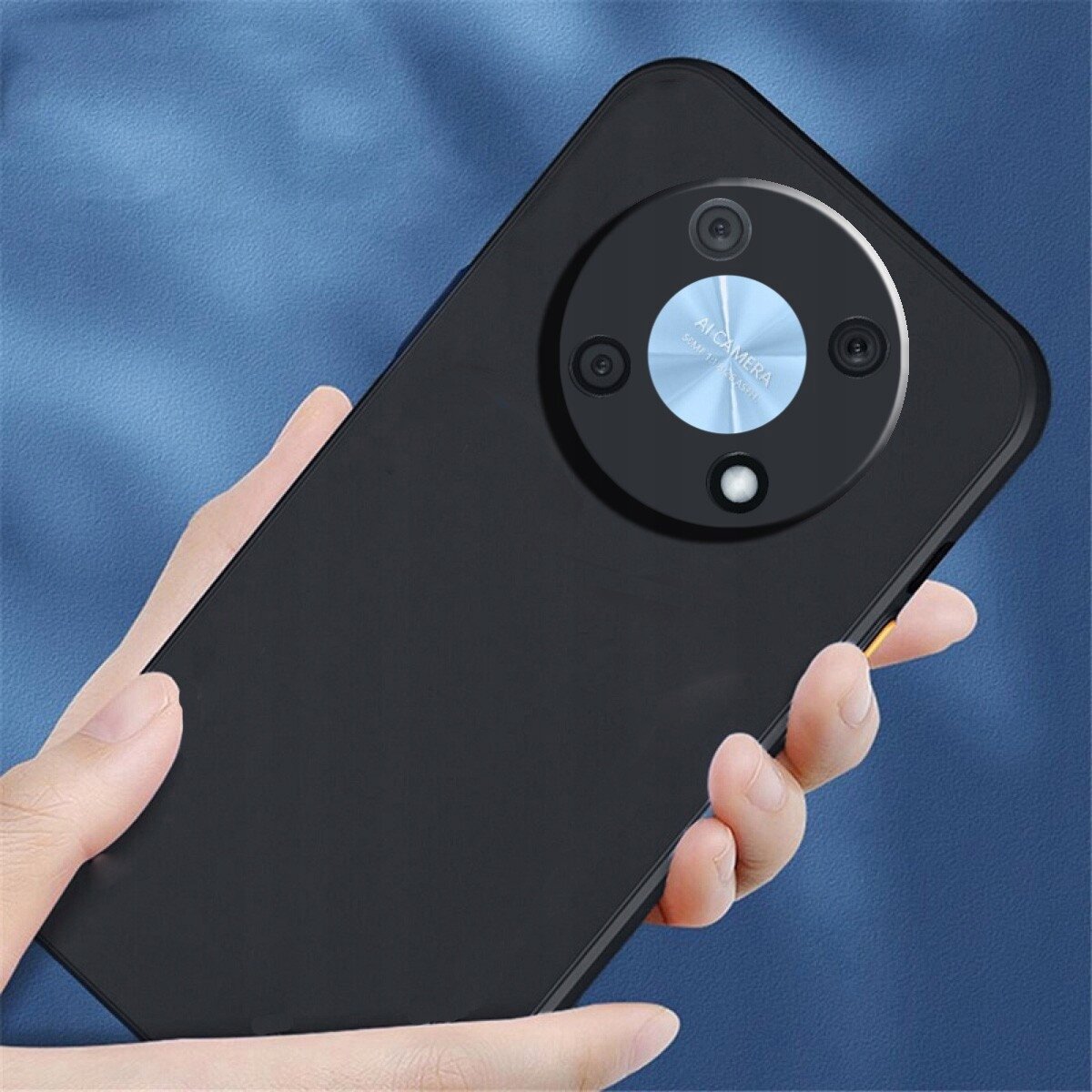 Czarne etui na telefon z okrągłym wycięciem na aparat, trzymane w dłoni na tle niebieskiego materiału. Na wycięciu widoczny napis \'AI CAMERA\'.