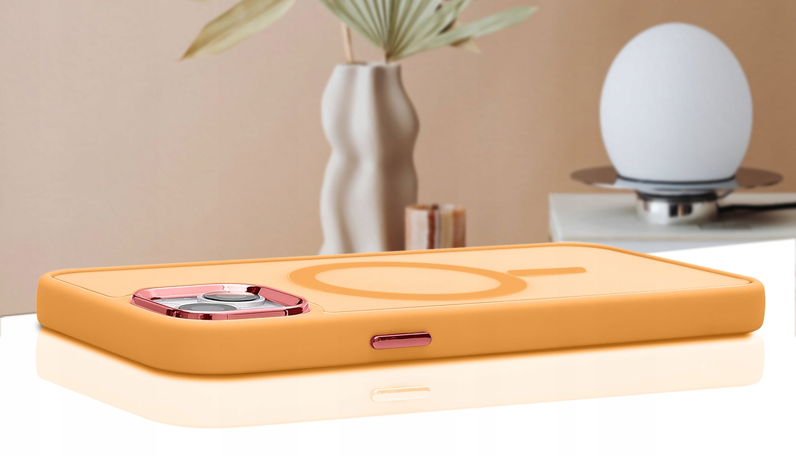 Pomarańczowe etui na telefon z trzema wycięciami na aparat, leży na białej powierzchni, w tle widoczny jest wazon z rośliną i lampa.
