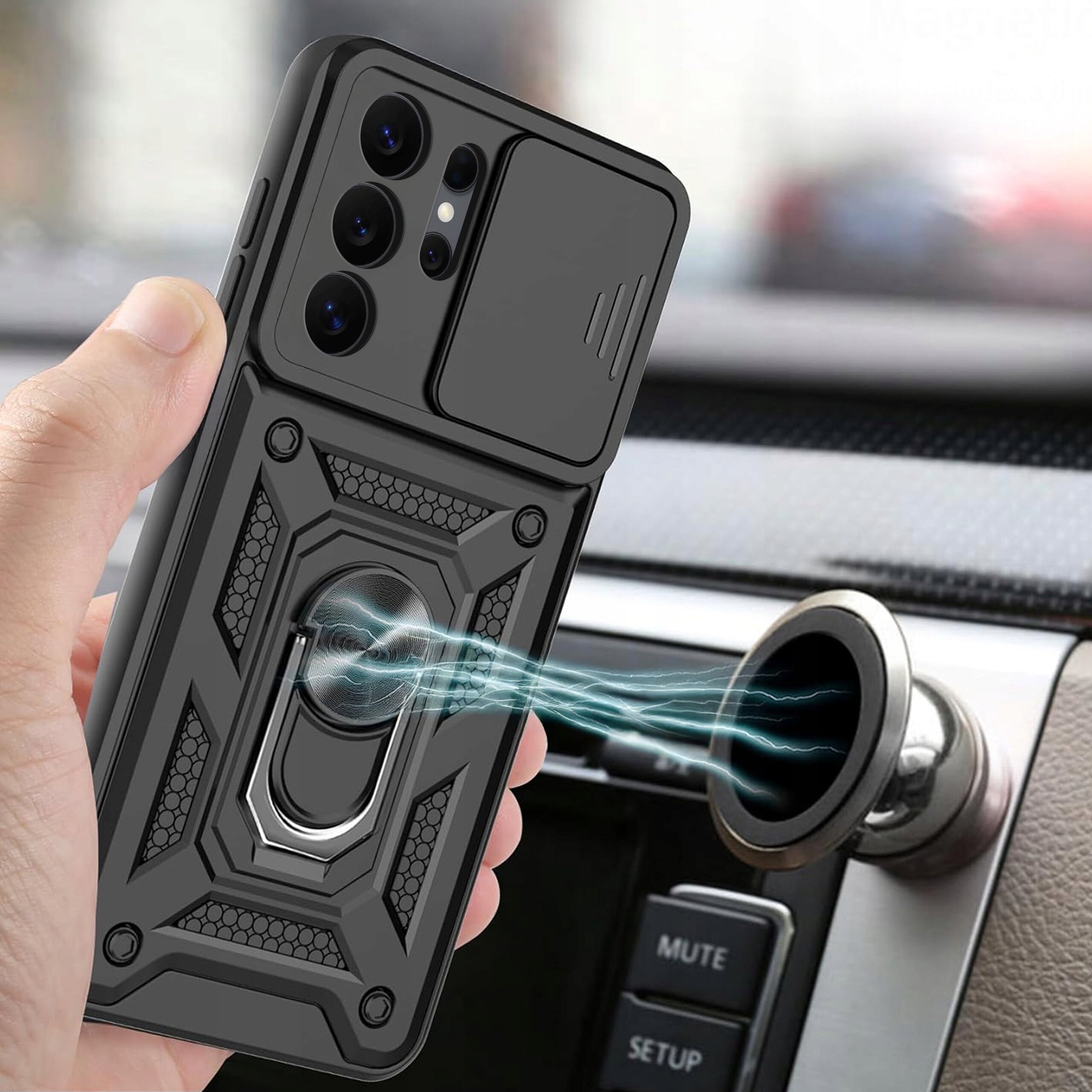 Osoba trzyma telefon w czarnym, pancernym etui z widocznym wzorem i metalowym uchwytem. W tle widać wnętrze samochodu z panelem przycisków, na którym znajduje się napis \'MUTE\' i \'SETUP\'.