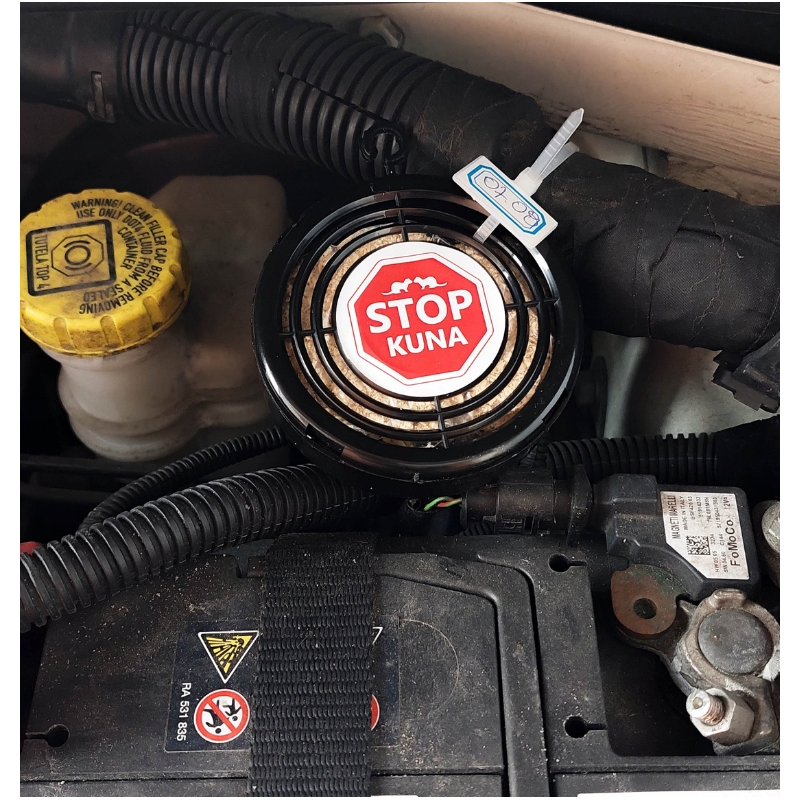 Na zdjęciu widać urządzenie z napisem \'STOP KUNA\' zamontowane w komorze silnika samochodu. Obok znajduje się żółty korek z ostrzeżeniem oraz przewody i elementy mechaniczne.