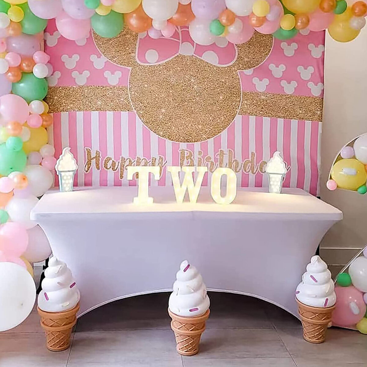 Dekoracja urodzinowa z balonami w pastelowych kolorach, tłem z różowymi i złotymi elementami oraz napisem \'Happy Birthday\'. Na stole znajduje się podświetlany napis \'TWO\' oraz dekoracje w kształcie lodów.