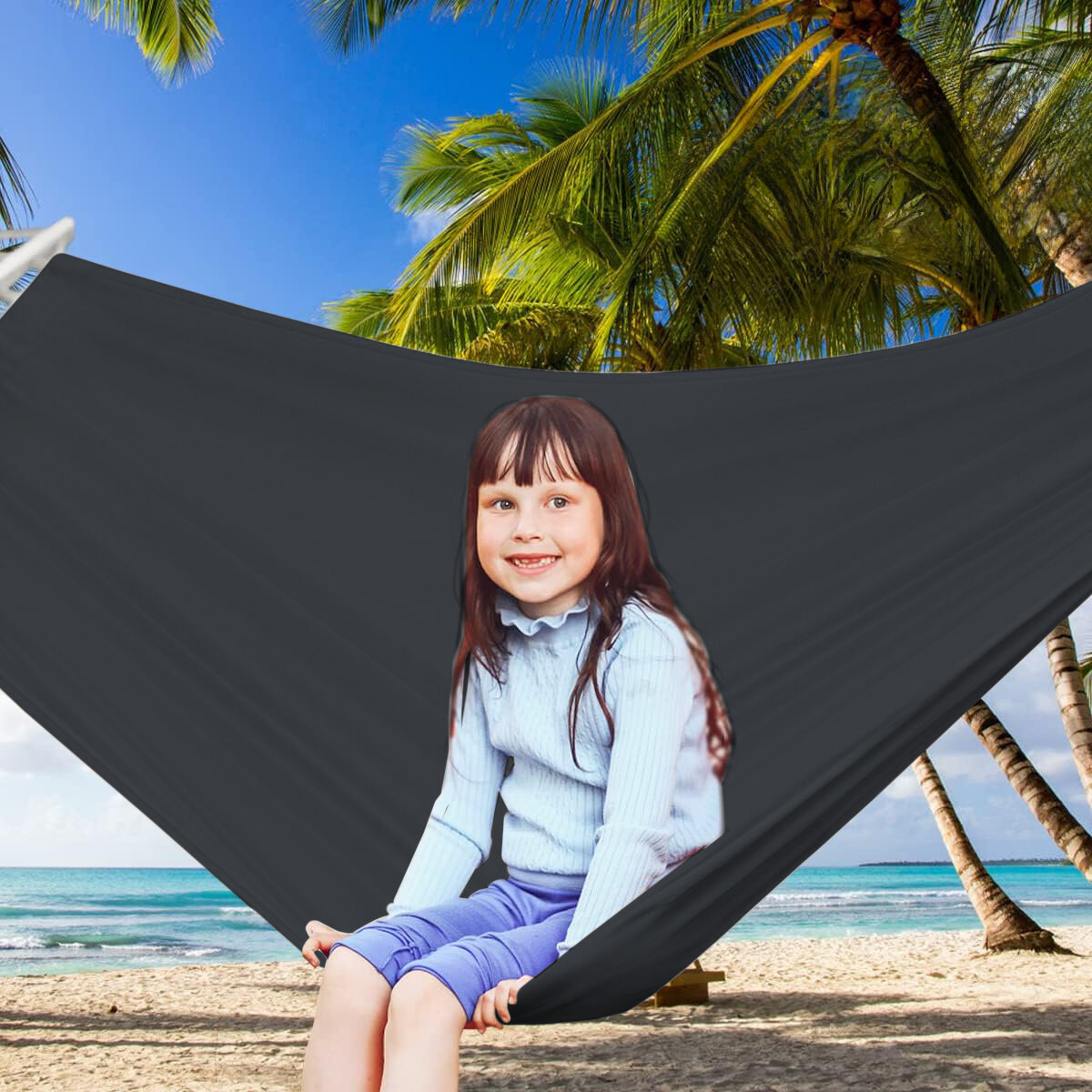 Grafitowy hamak sznurkowo-drewniany zawieszony na plaży z palmami w tle. Na hamaku siedzi dziecko ubrane w jasną bluzkę i niebieskie spodnie.