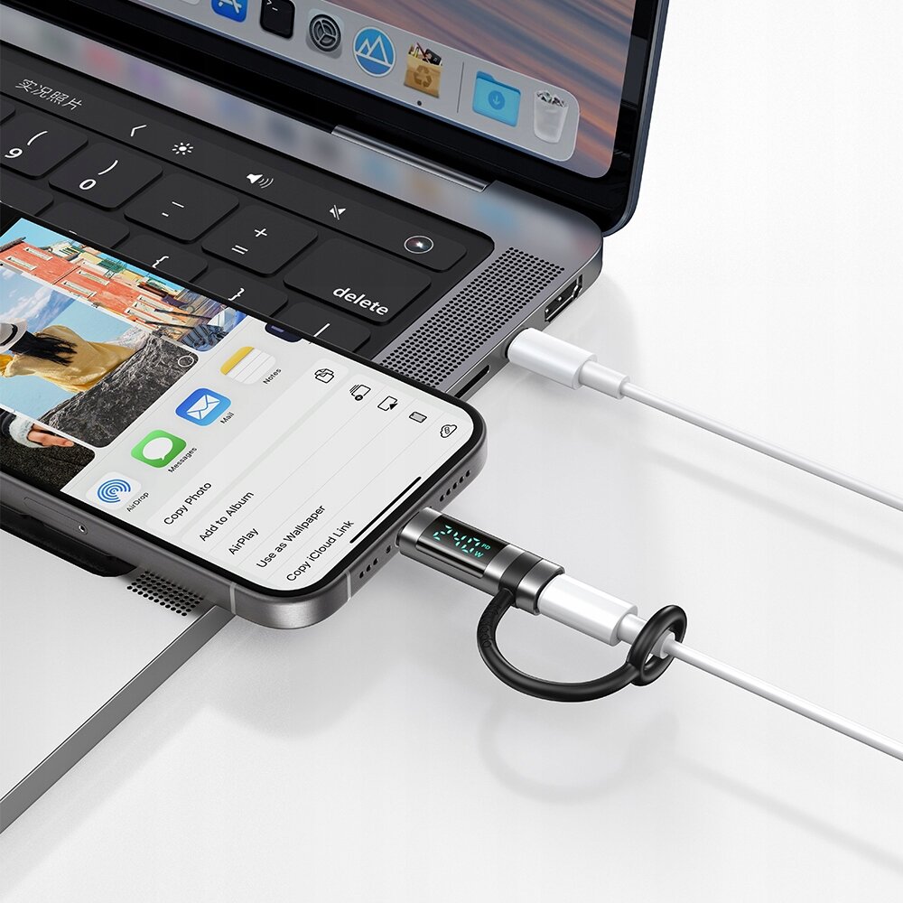 Zdjęcie przedstawia laptop z otwartym ekranem i klawiaturą, do którego podłączony jest smartfon za pomocą adaptera USB-C do Lightning. Na ekranie smartfona widoczne są ikony aplikacji i opcje udostępniania zdjęcia.