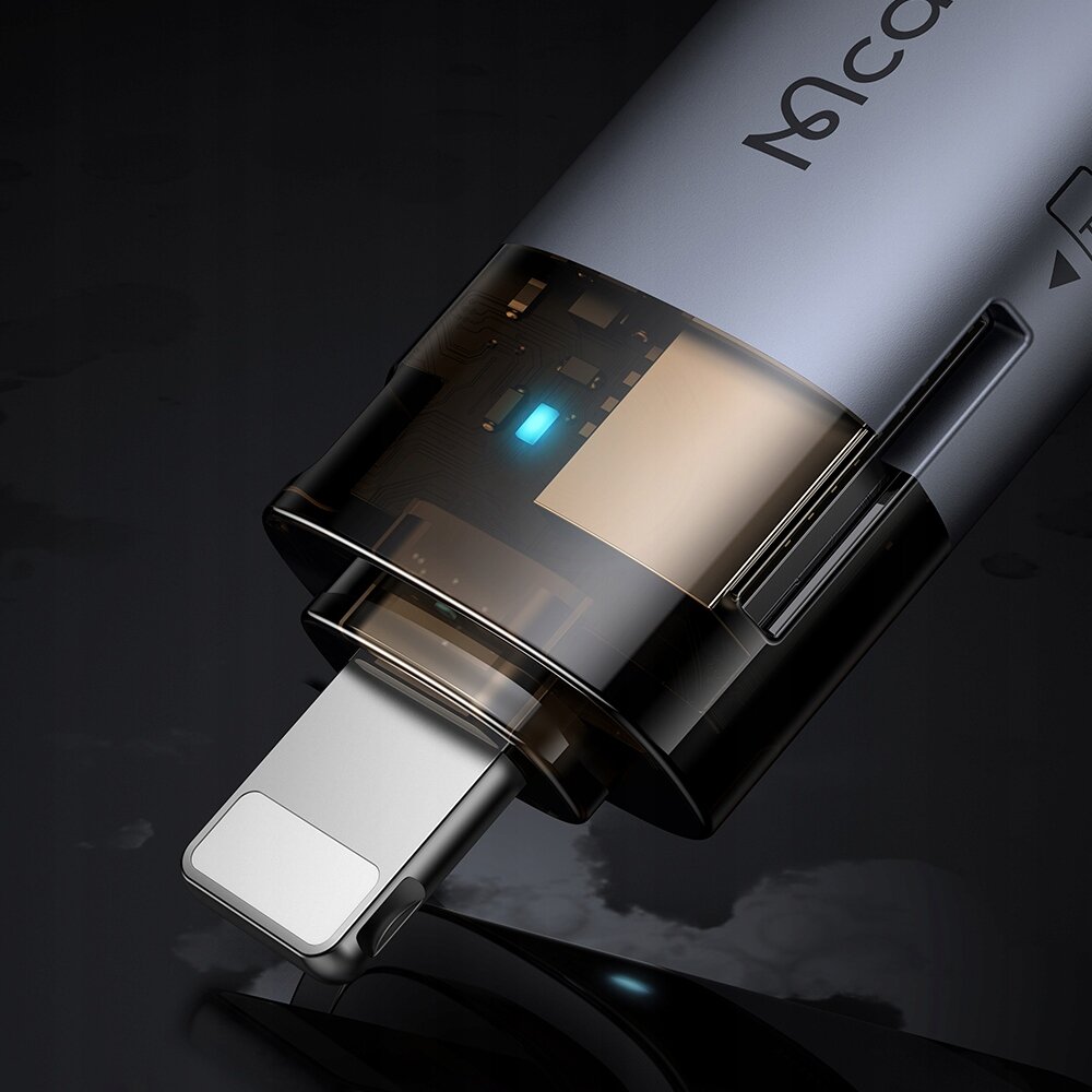 Zbliżenie na adapter USB-A do Lightning z widocznym logo \'Mcdodo\' na obudowie oraz podświetlonym elementem wewnętrznym. Produkt ma metaliczny wygląd i przezroczystą część obudowy, przez którą widać wewnętrzne komponenty.