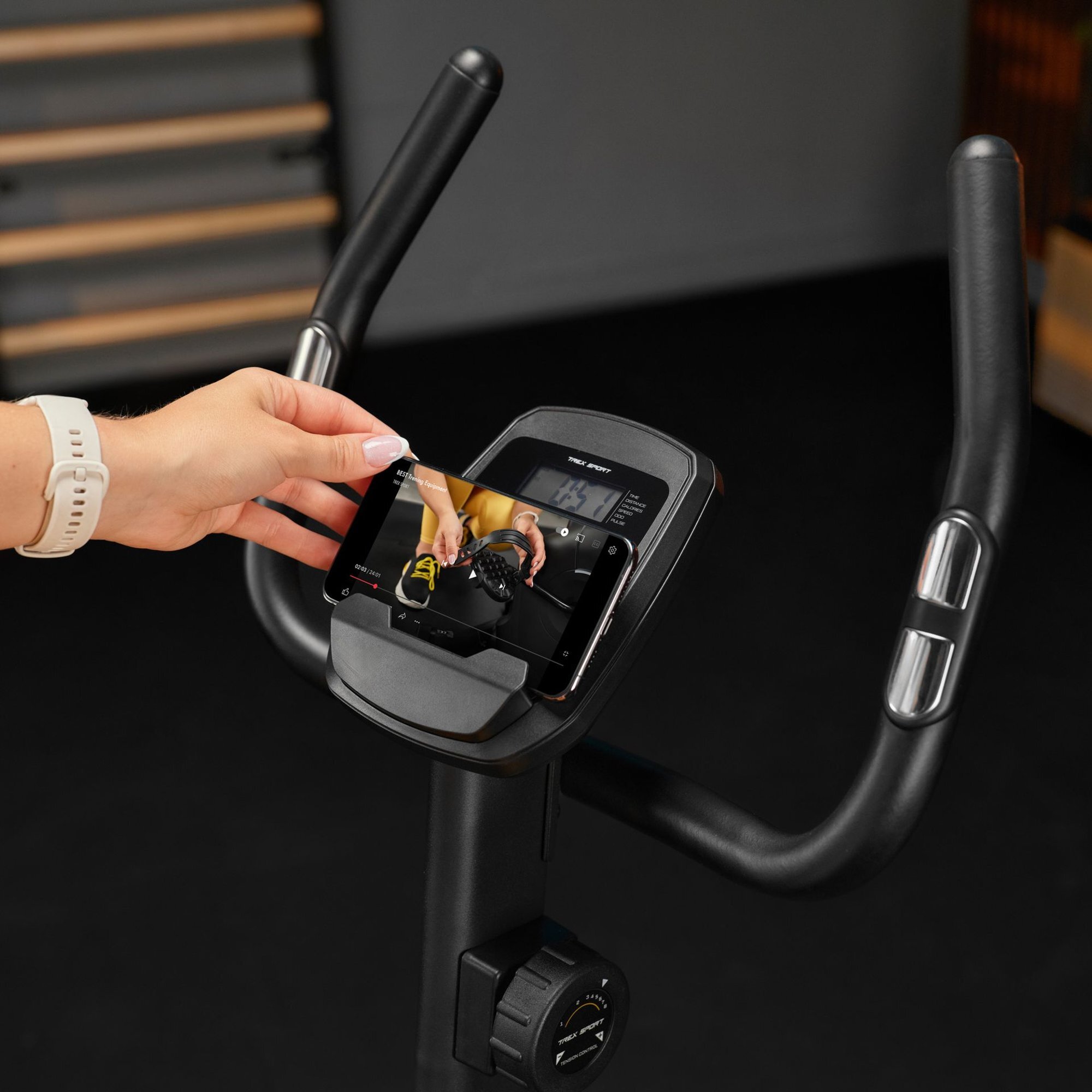 Osoba trzyma smartfon na uchwycie roweru stacjonarnego, na ekranie widoczny jest film instruktażowy. Rower posiada czarną kierownicę z wyświetlaczem i pokrętłem regulacji oporu.