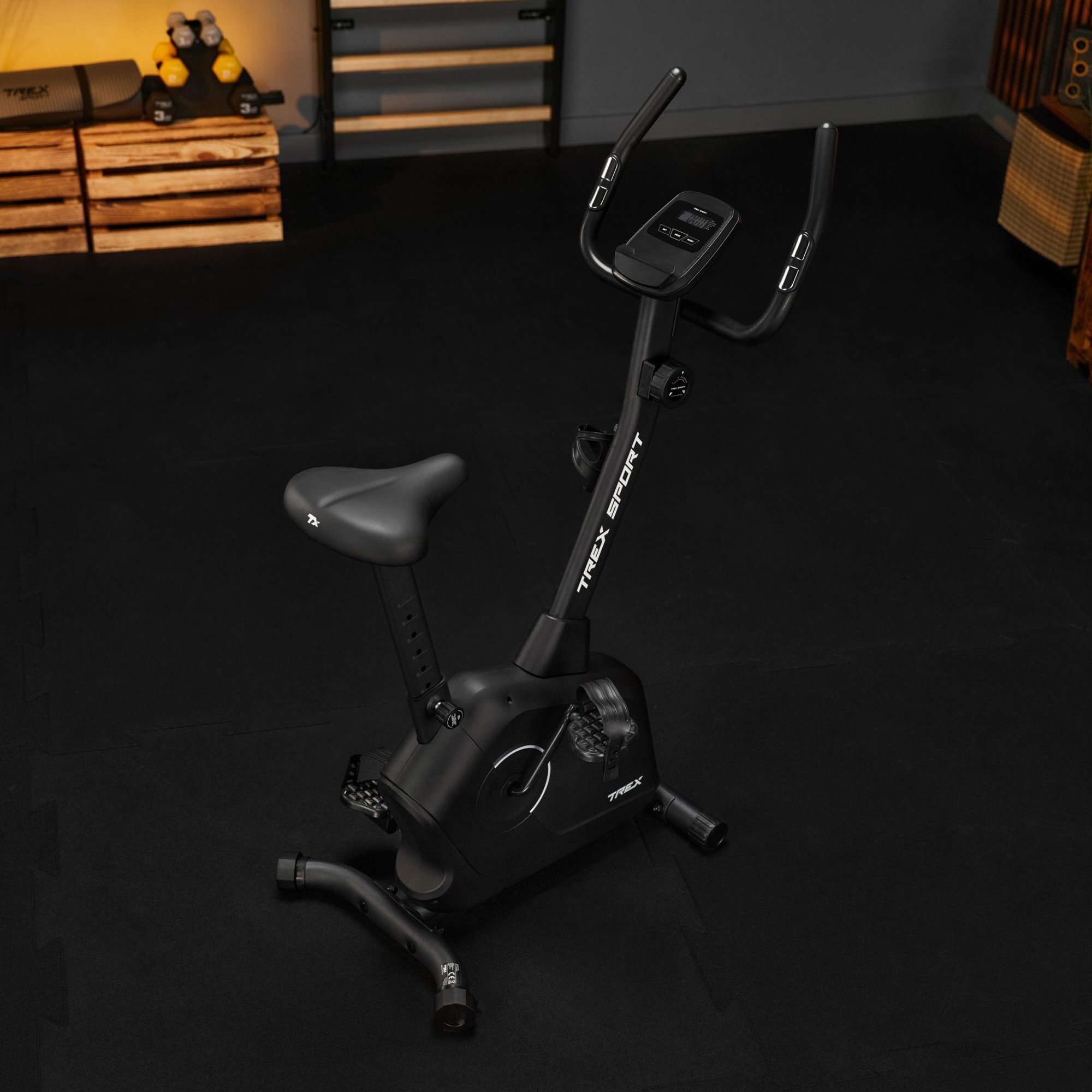 Czarny rower stacjonarny z wyświetlaczem i ergonomicznym siodełkiem, na ramie widoczny napis \'TREX SPORT\'. W tle znajdują się drewniane skrzynki i sprzęt do ćwiczeń.