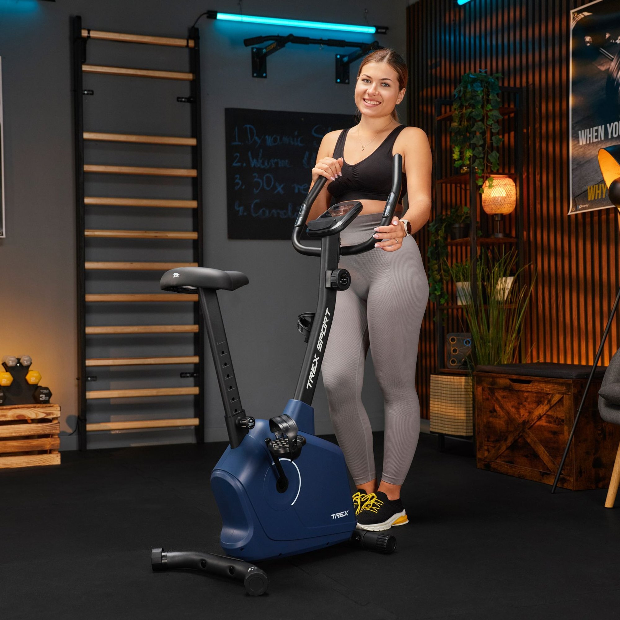 Osoba stoi obok roweru stacjonarnego magnetycznego w kolorze granatowym z napisem \'TREK SPORT\'. W tle widoczne są drabinki gimnastyczne, tablica z napisami oraz rośliny.