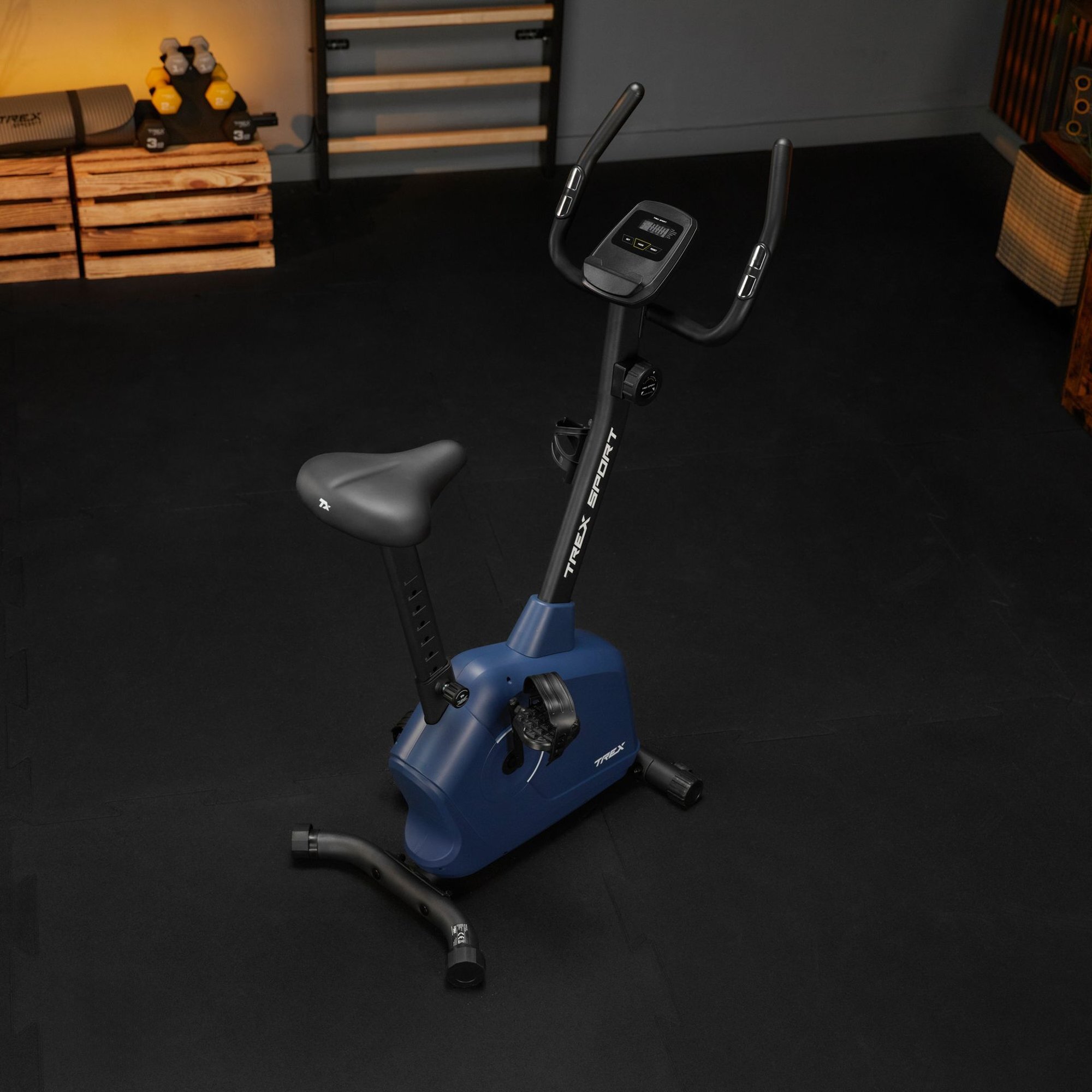 Na obrazie znajduje się rower stacjonarny w kolorze granatowym z czarnym siodełkiem i kierownicą. Na ramie widoczny jest napis \'TREX SPORT\'.