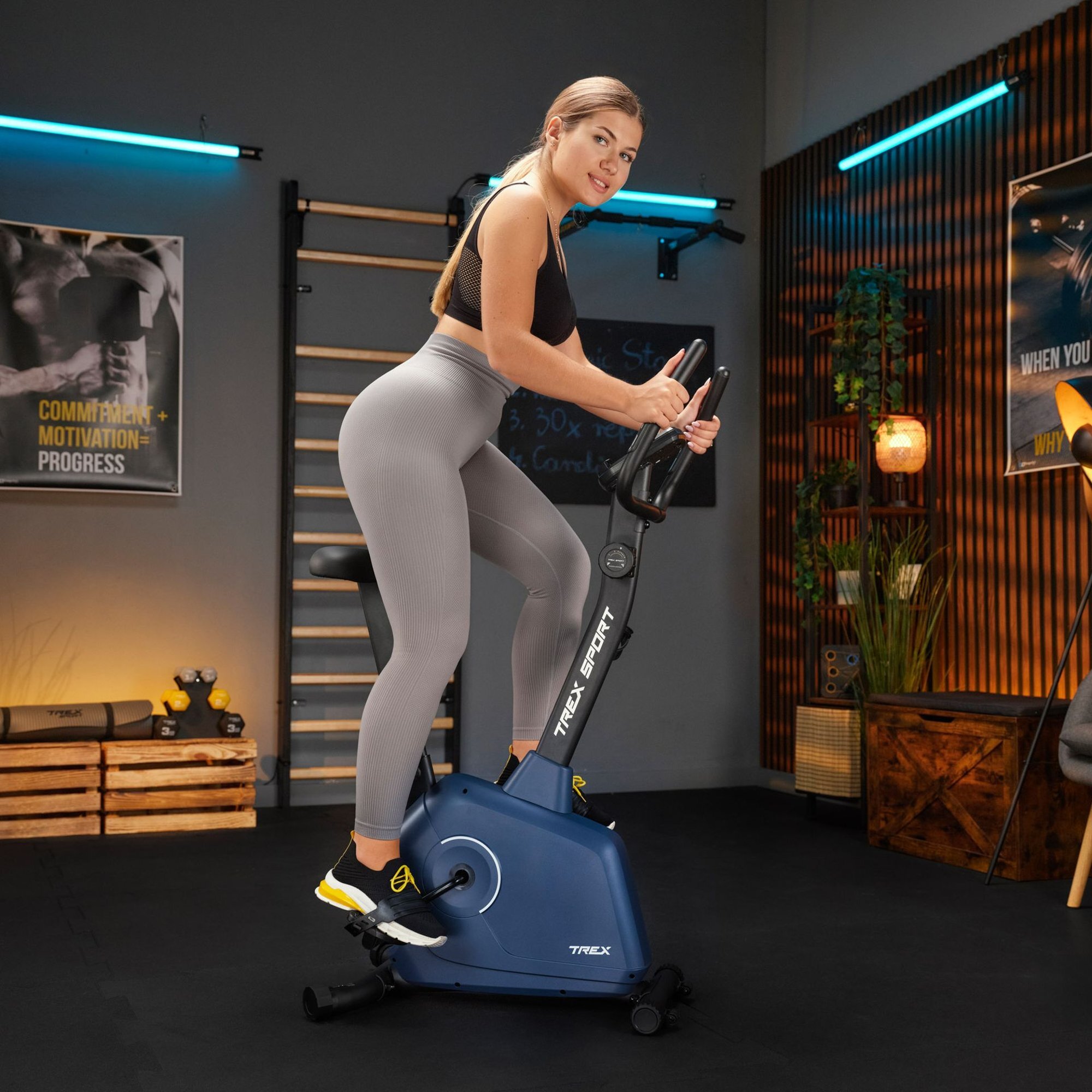 Osoba ćwiczy na rowerze stacjonarnym w pomieszczeniu z drewnianymi elementami i plakatami na ścianach. Rower ma napis \'TREX SPORT\' i jest w kolorze granatowym.