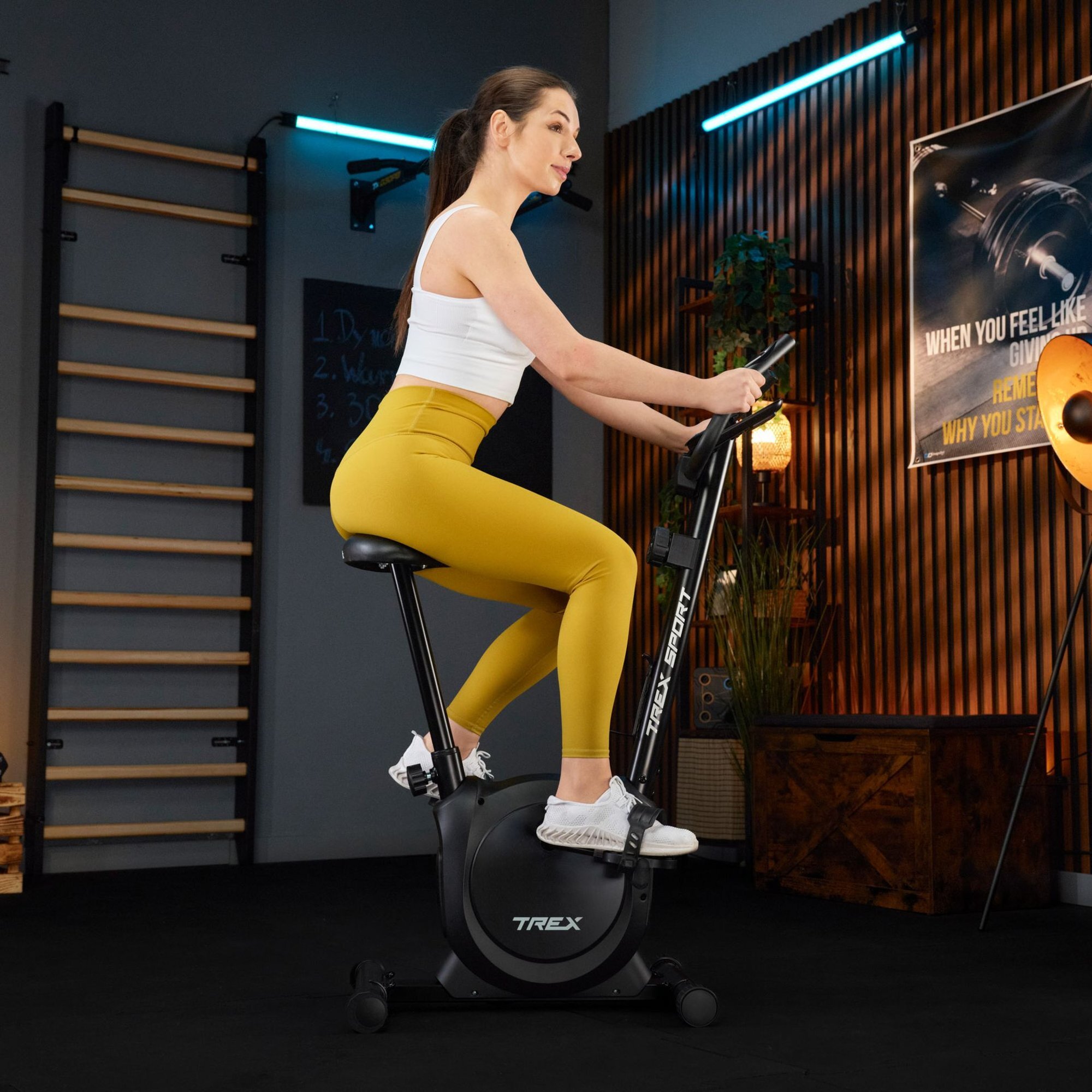 Osoba ćwiczy na czarnym rowerze stacjonarnym w pomieszczeniu z drewnianymi elementami dekoracyjnymi. Na rowerze widoczne jest logo \'TREX\'.