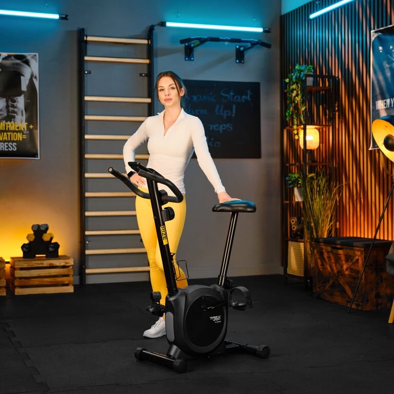 Kobieta stoi obok czarnego roweru stacjonarnego magnetycznego RIZE TX-350MB w pomieszczeniu z drewnianymi elementami dekoracyjnymi. Na ścianie widoczna jest tablica z napisem \'Start Warm Up!\'.